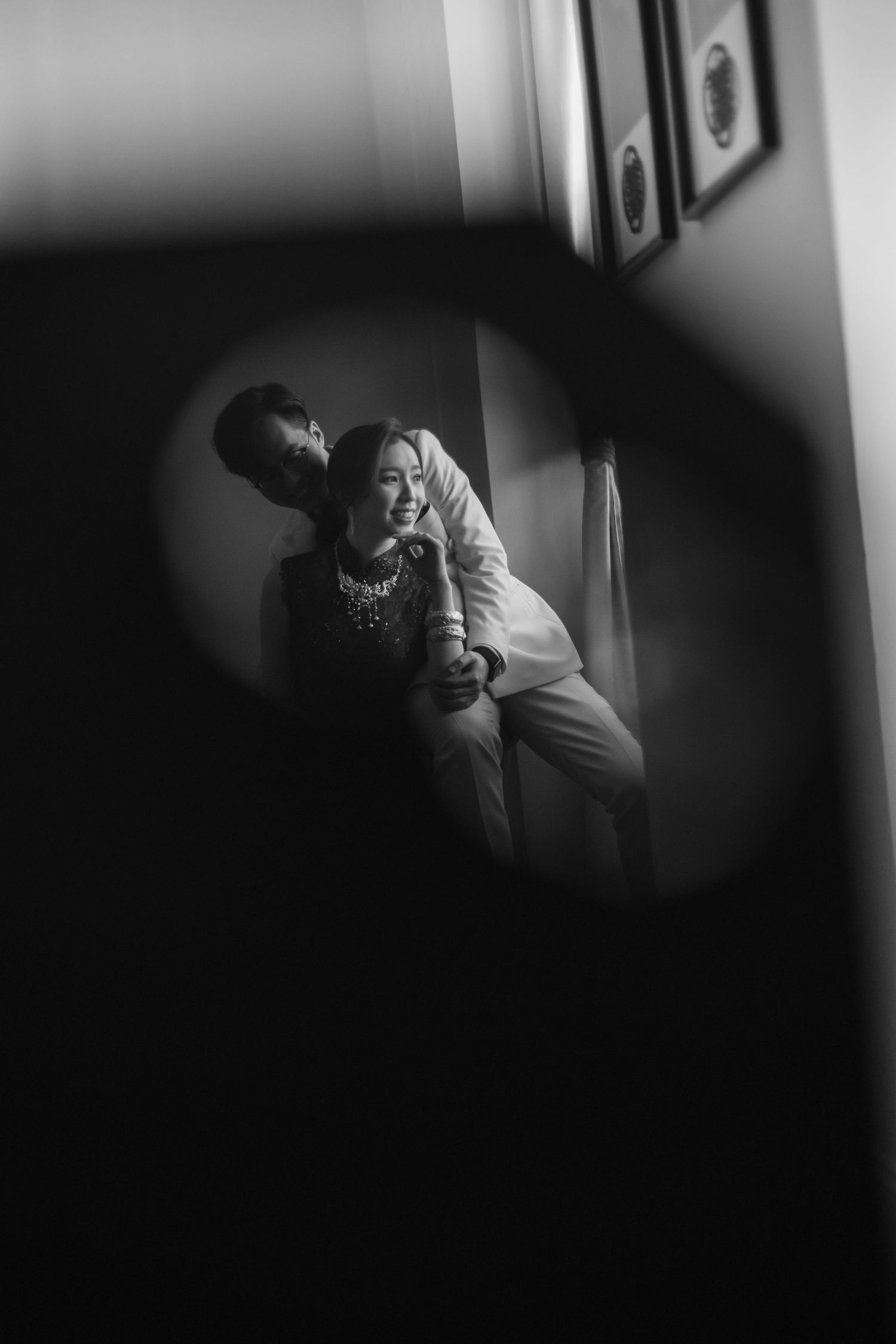 Black and white romantic portrait of couple embracing by window reflected in circular mirror.