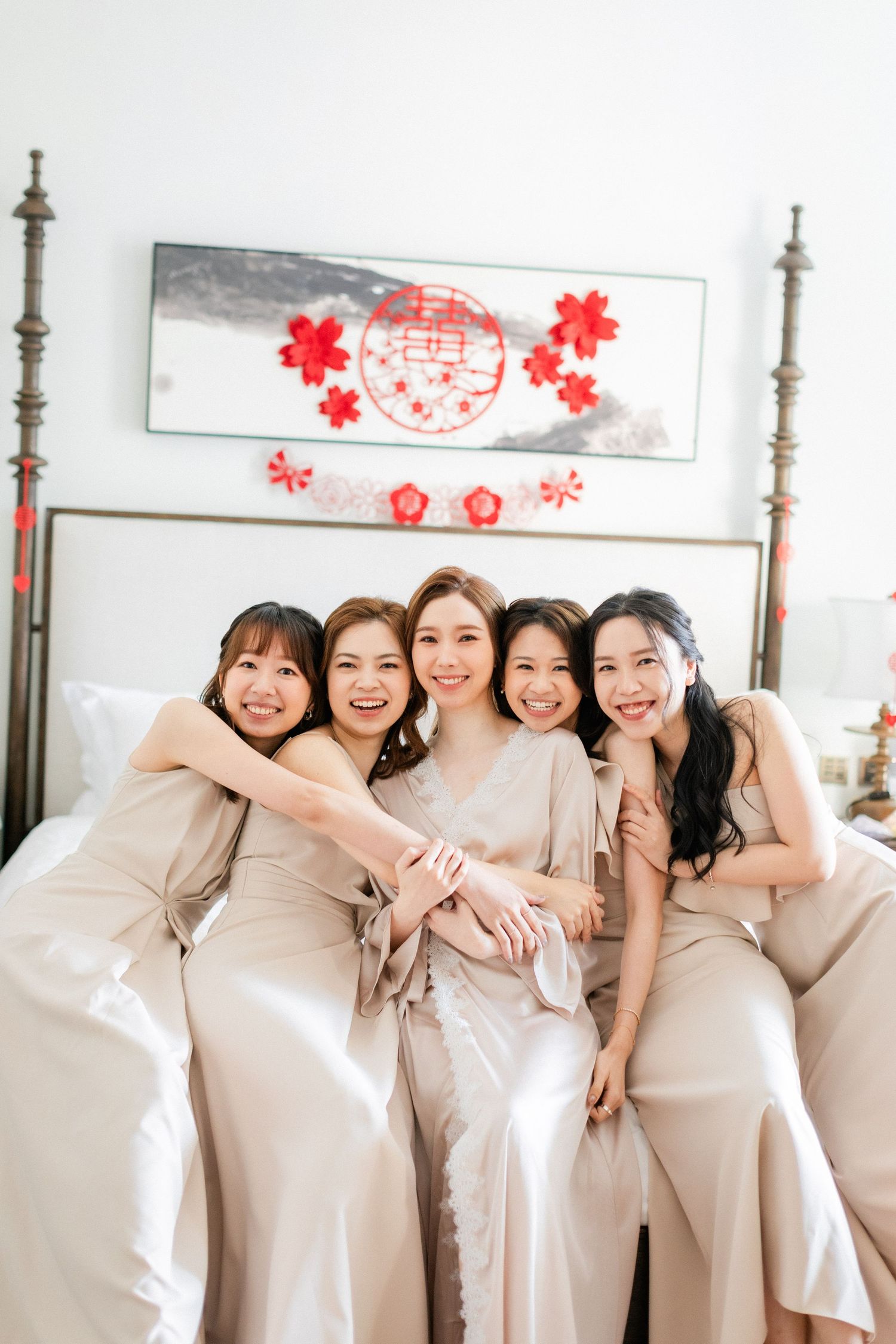 A bride in a white robe sits on a bed with four bridesmaids in champagne satin dresses, all smiling and embracing.