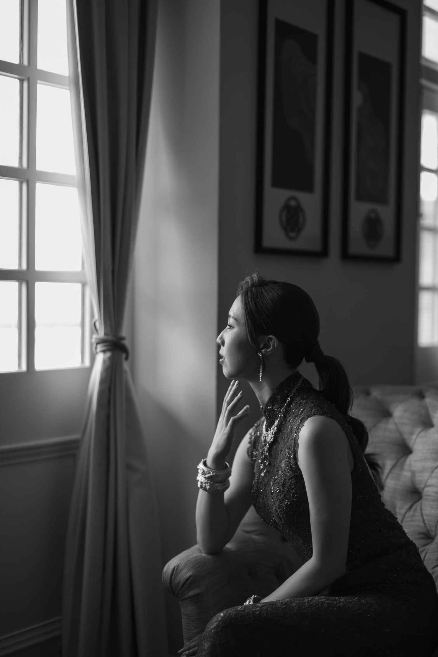 Elegant bride in black dress gazes pensively out window in moody black and white portrait.