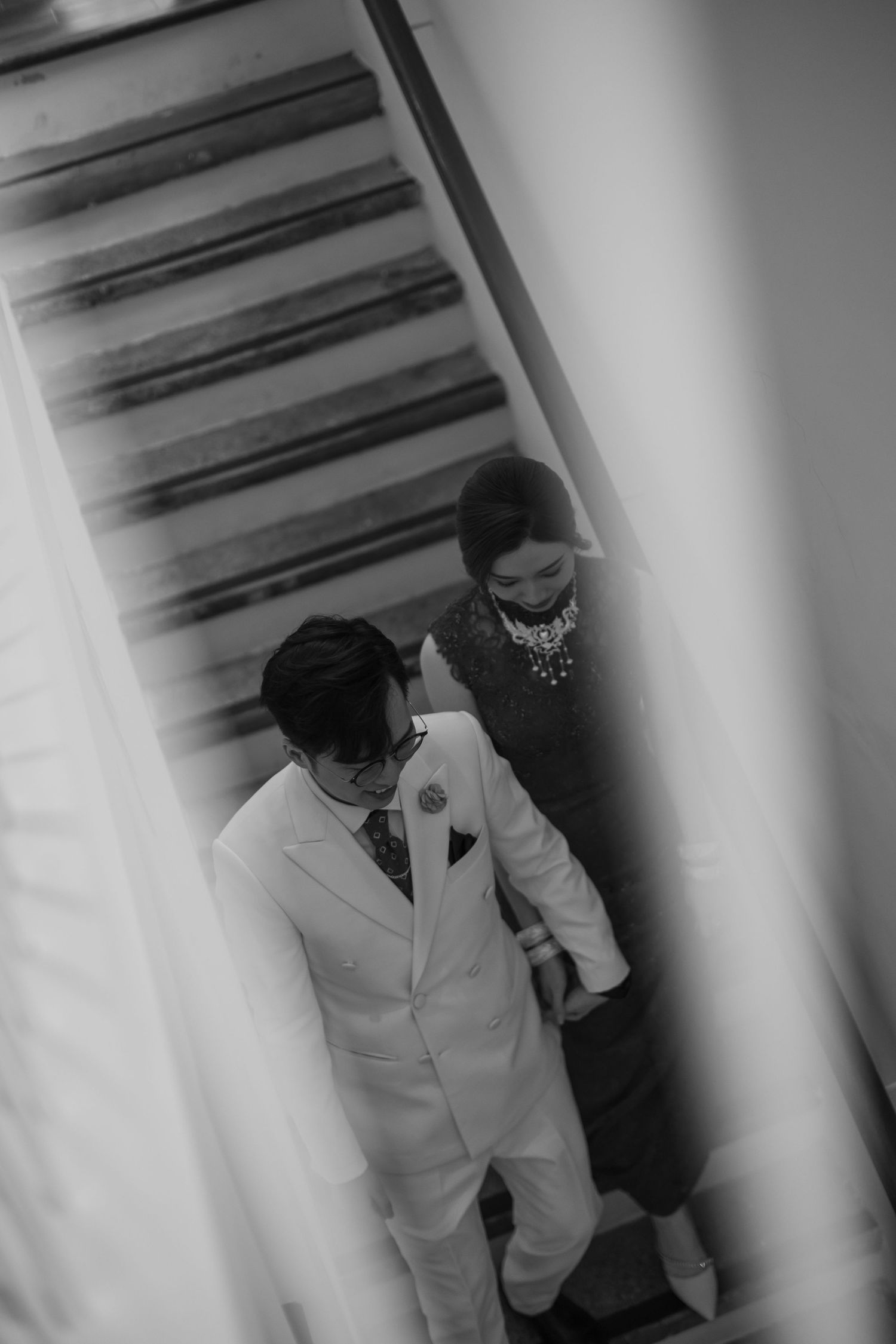 Black and white overhead shot of groom in white suit and bride descending staircase together.
