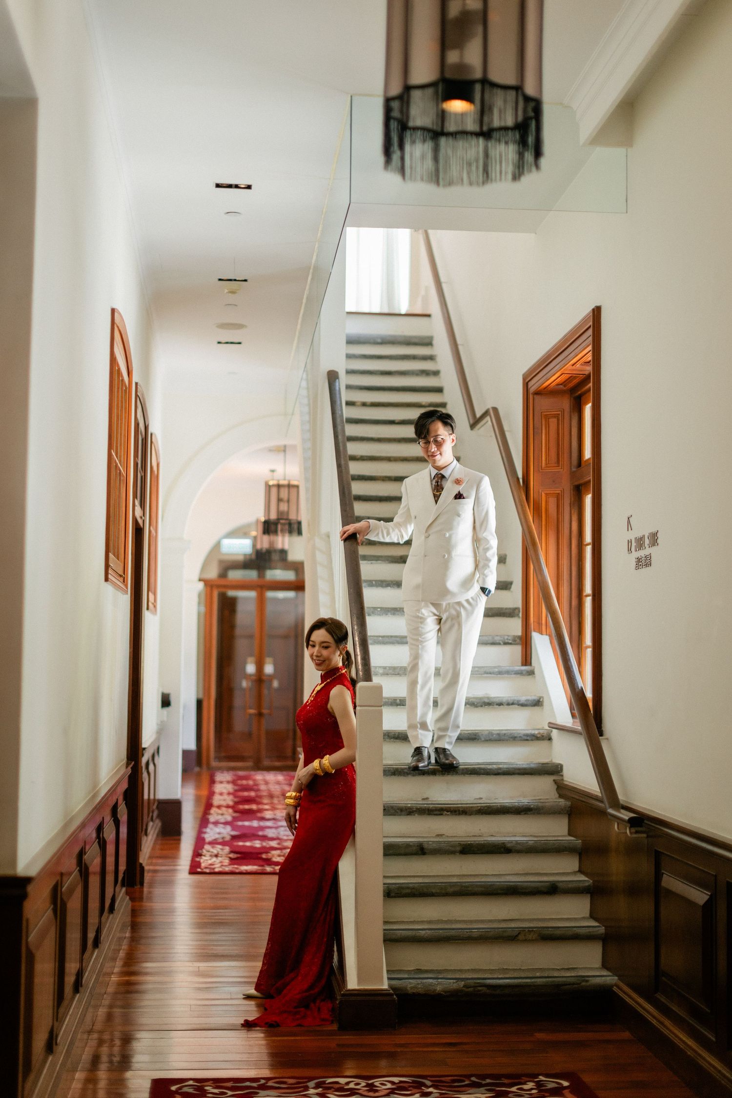 Bride in red qipao and groom in white suit pose elegantly on grand staircase in historic venue.