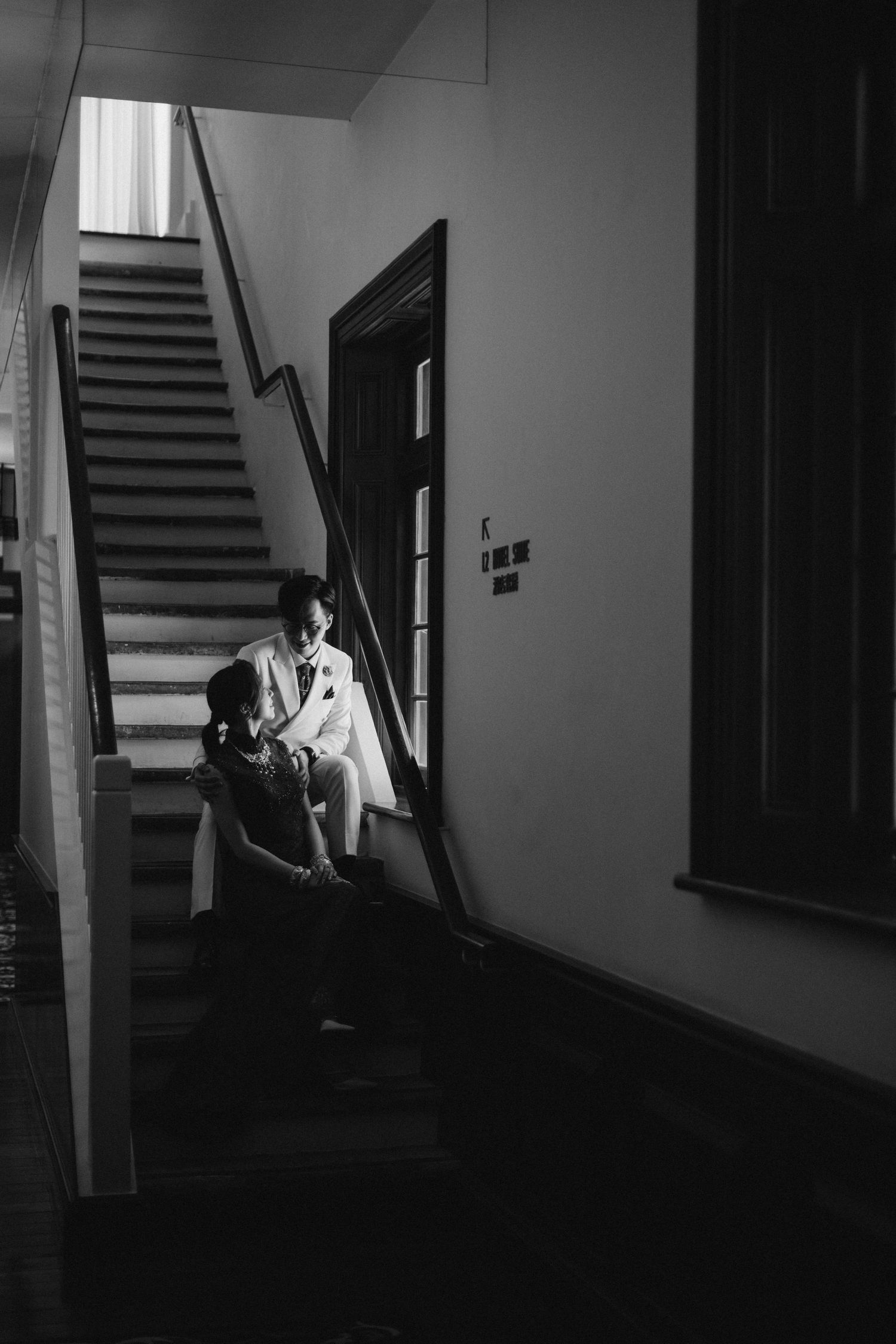 Black and white artistic photo of couple sitting together on staircase steps in dramatic light.