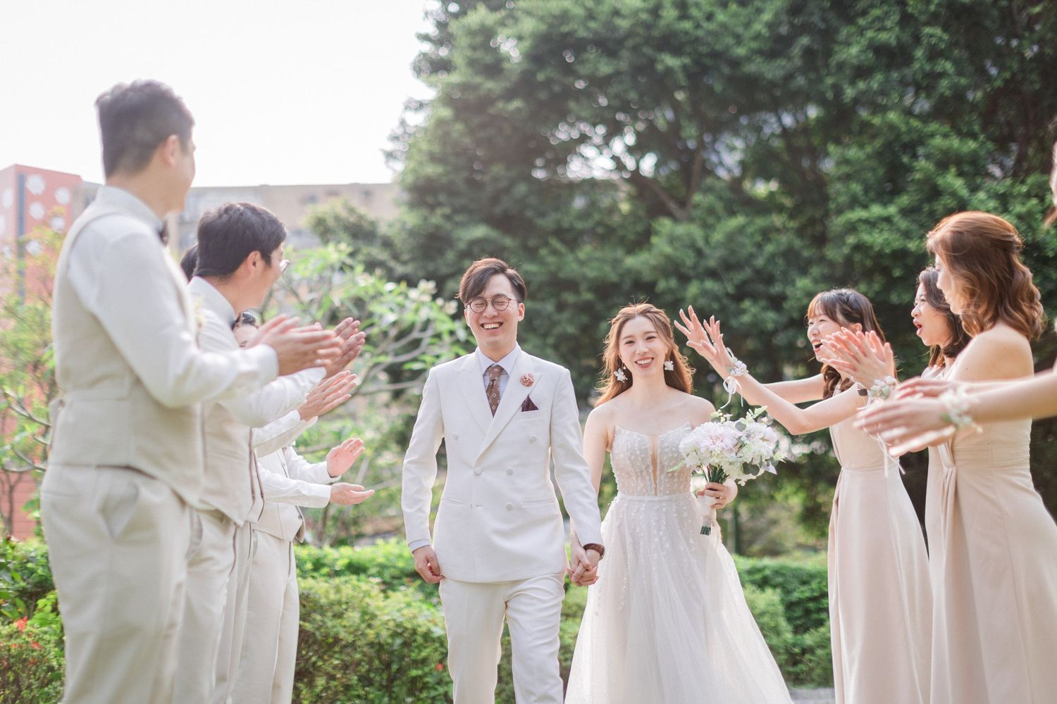 Bride in white gown and groom in white suit walk through cheering wedding party outdoors.