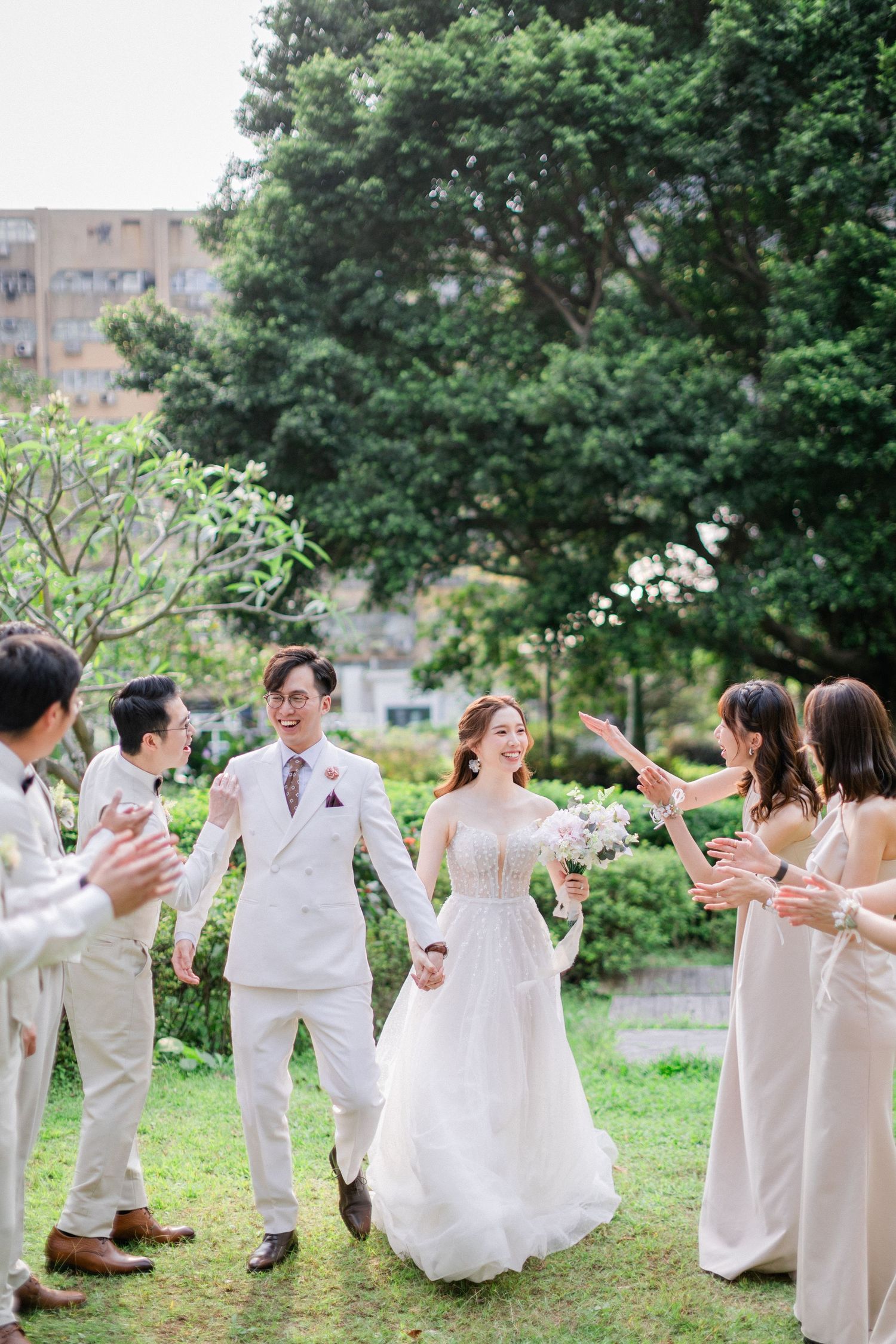 Bride and groom walk hand-in-hand through a garden aisle as wedding party guests applaud in celebration.