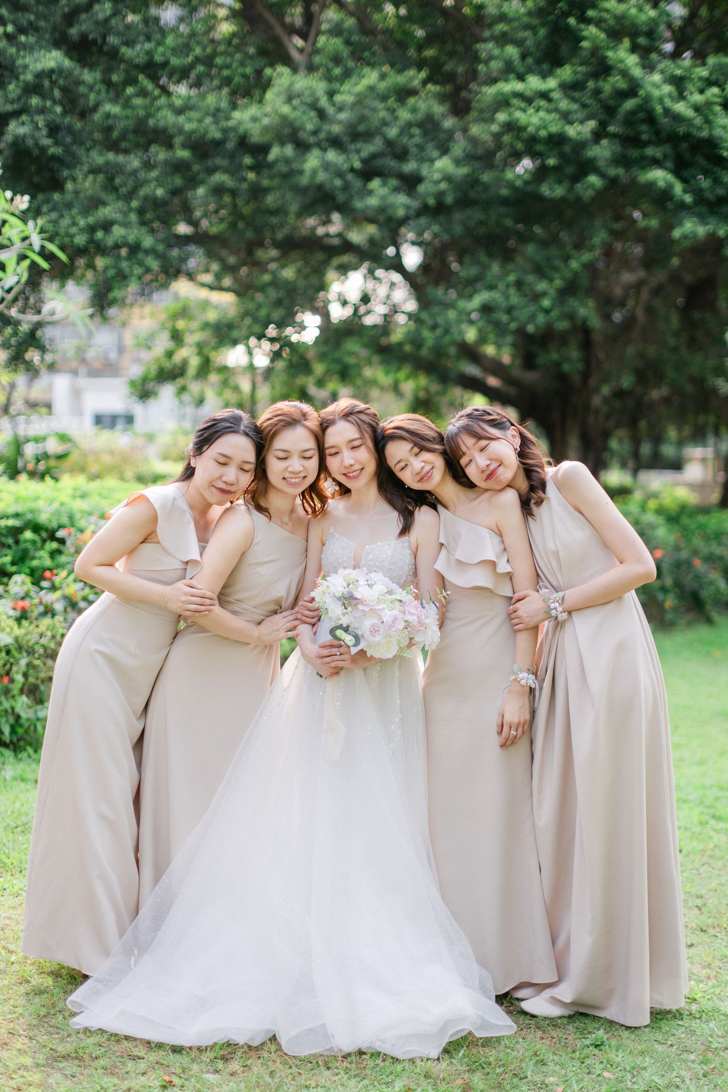 Bride in white gown holds bouquet surrounded by four bridesmaids in beige dresses in a lush garden.