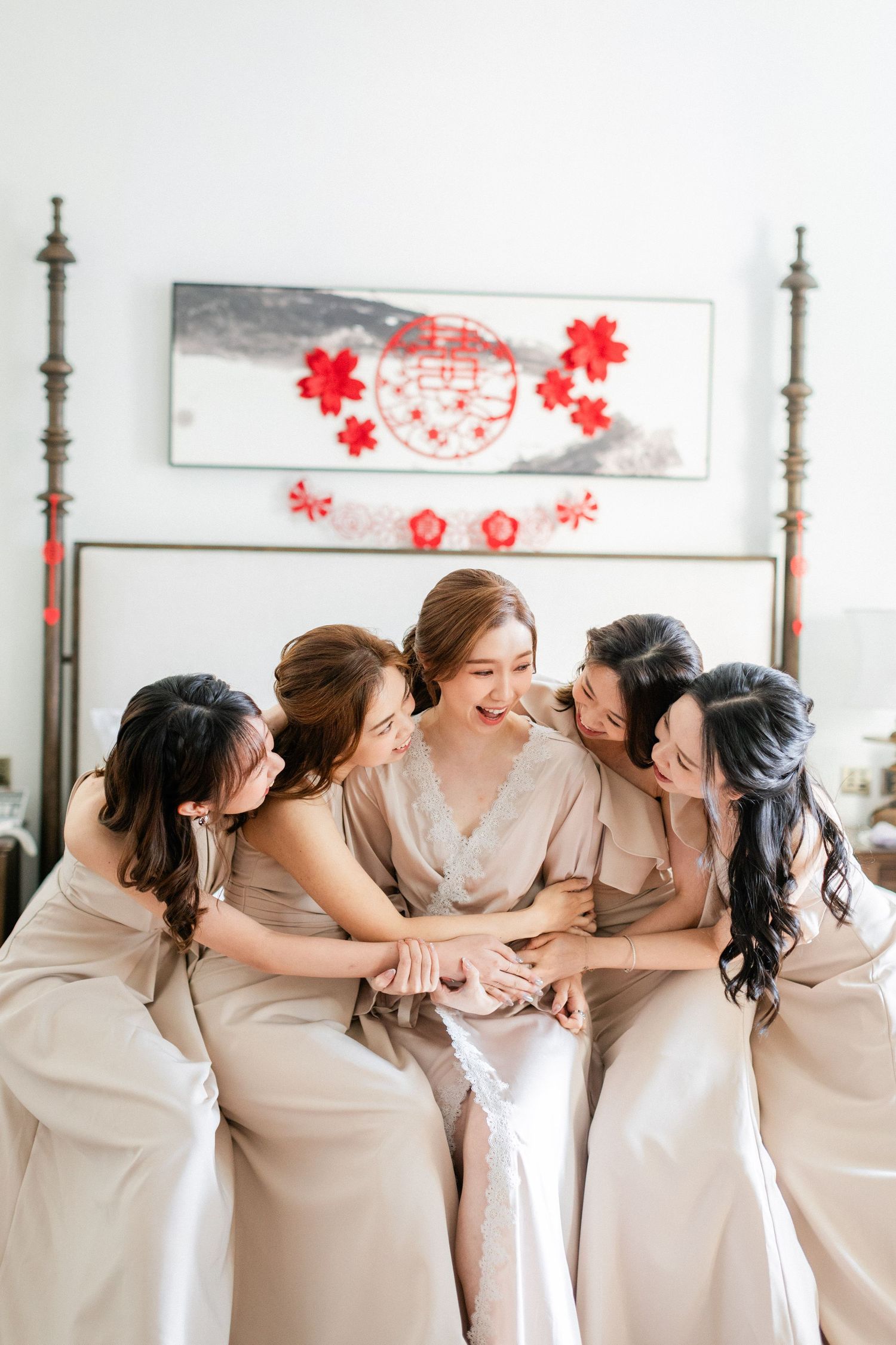 A bride in a white robe laughs joyfully as four bridesmaids in champagne dresses huddle close together on a bed.