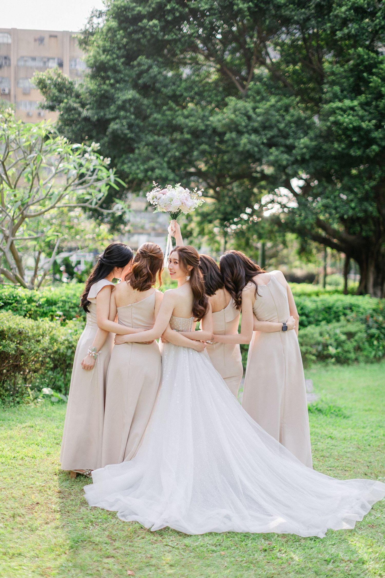 Bride and four bridesmaids in beige gowns share a tender group hug from behind in a green garden.