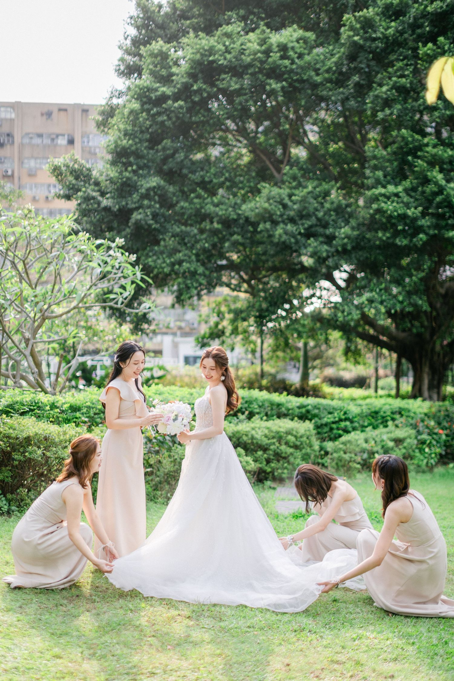 Bridesmaids in beige dresses help arrange the bride's white wedding gown train on a garden lawn.