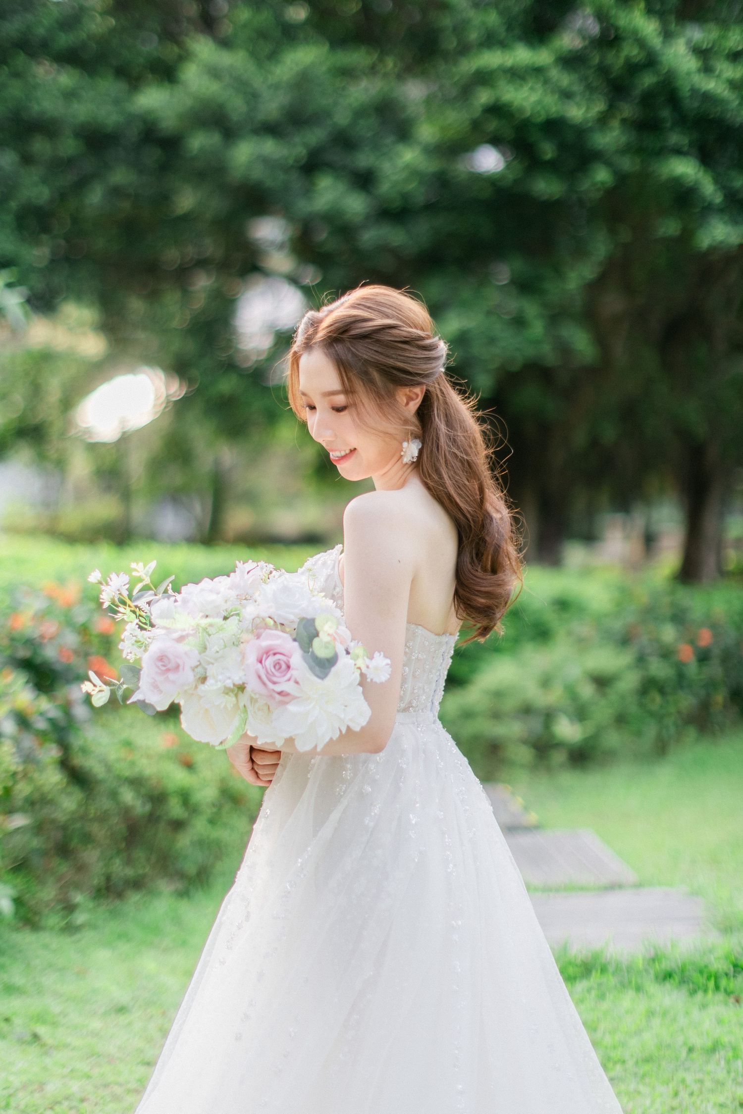 Elegant bride in a white lace gown holds a soft pink and white floral bouquet in a sunlit garden.