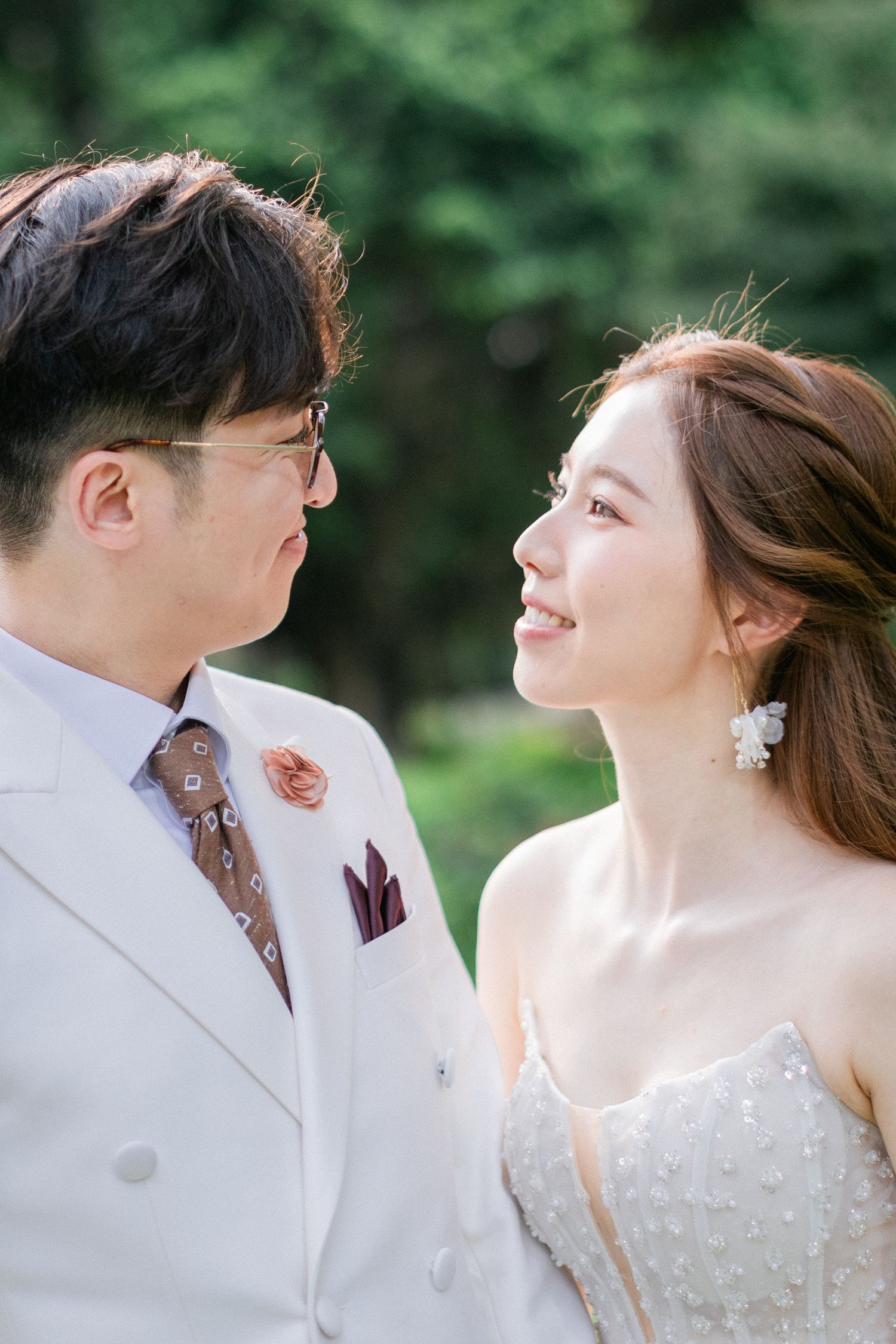Groom in white suit and bride in white gown share a romantic gaze outdoors in soft natural light.