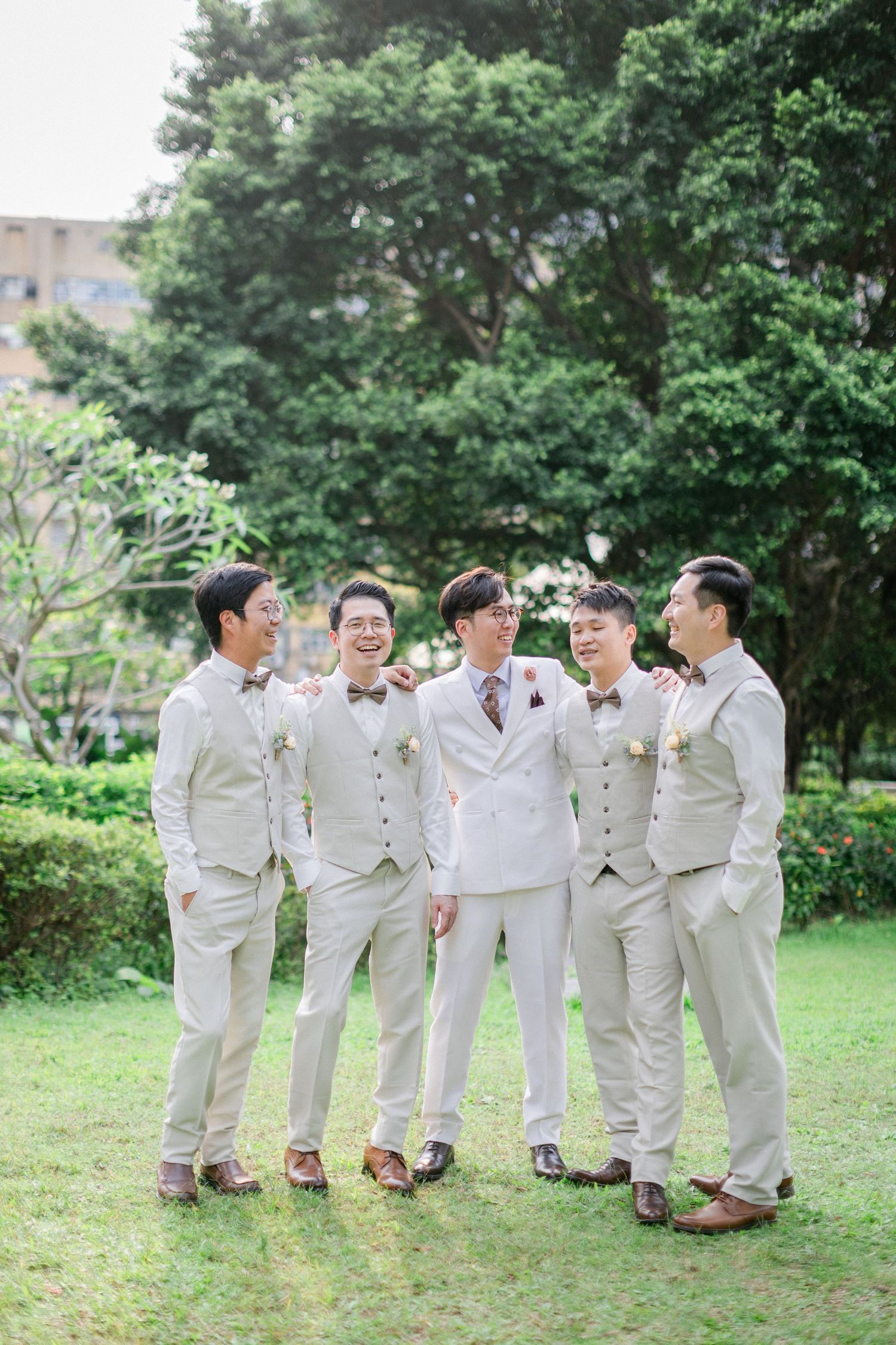 Groom in white double-breasted suit stands smiling with four groomsmen in matching beige suits outdoors.