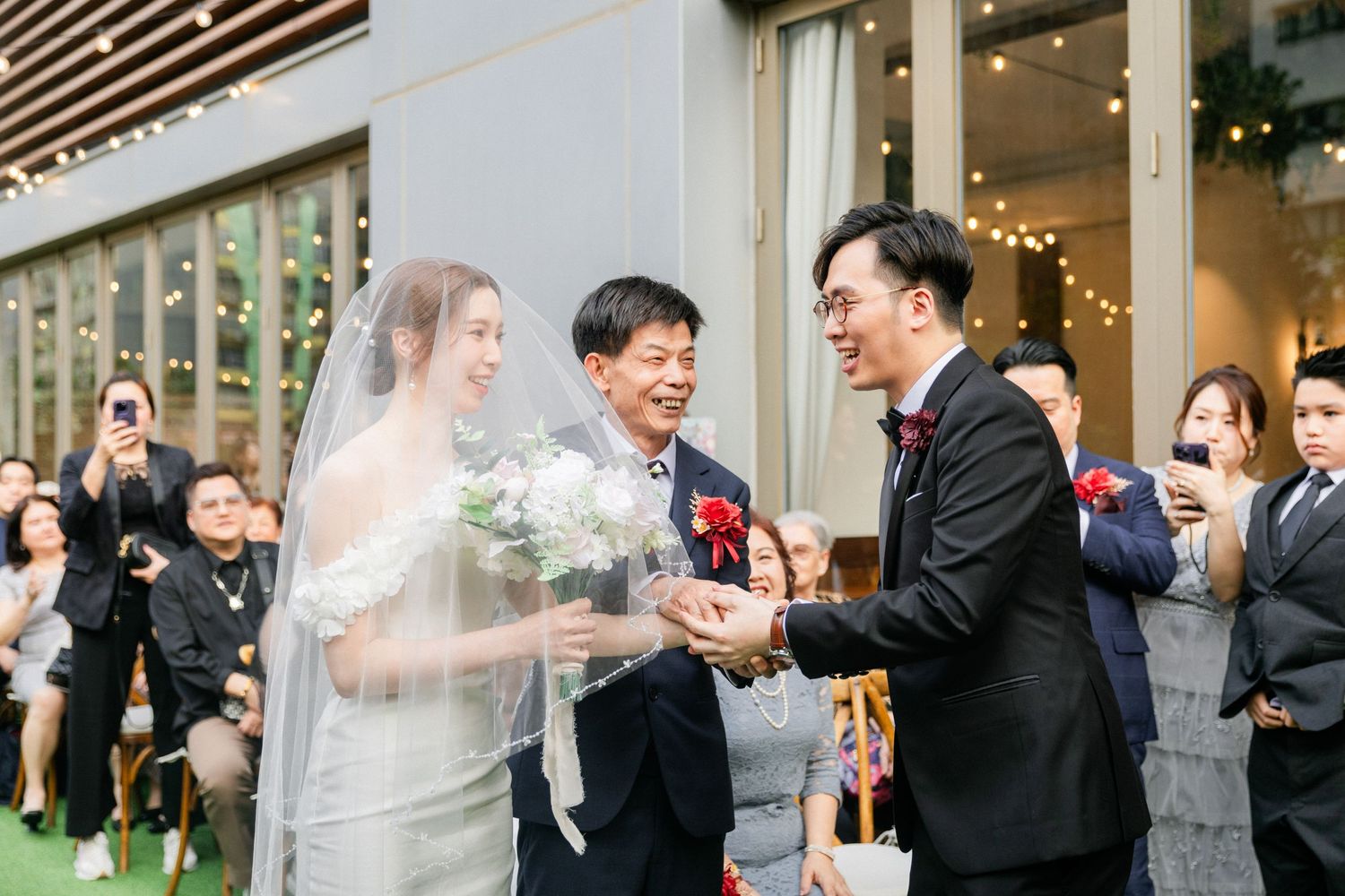 Bride in white veil receives a joyful handshake during an outdoor wedding ceremony surrounded by guests.