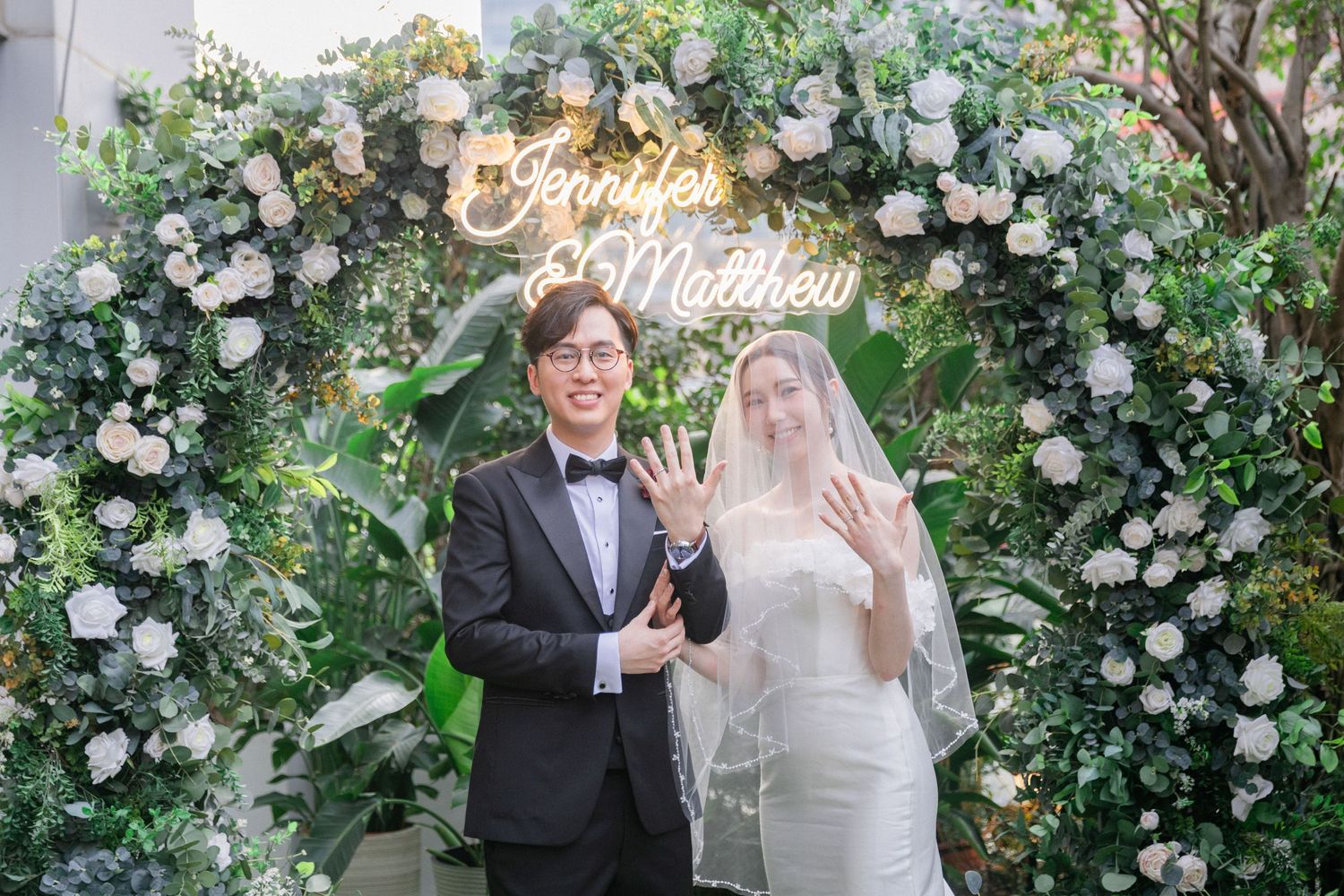Newlyweds Jennifer and Matthew pose showing wedding rings before a floral arch with a neon name sign.