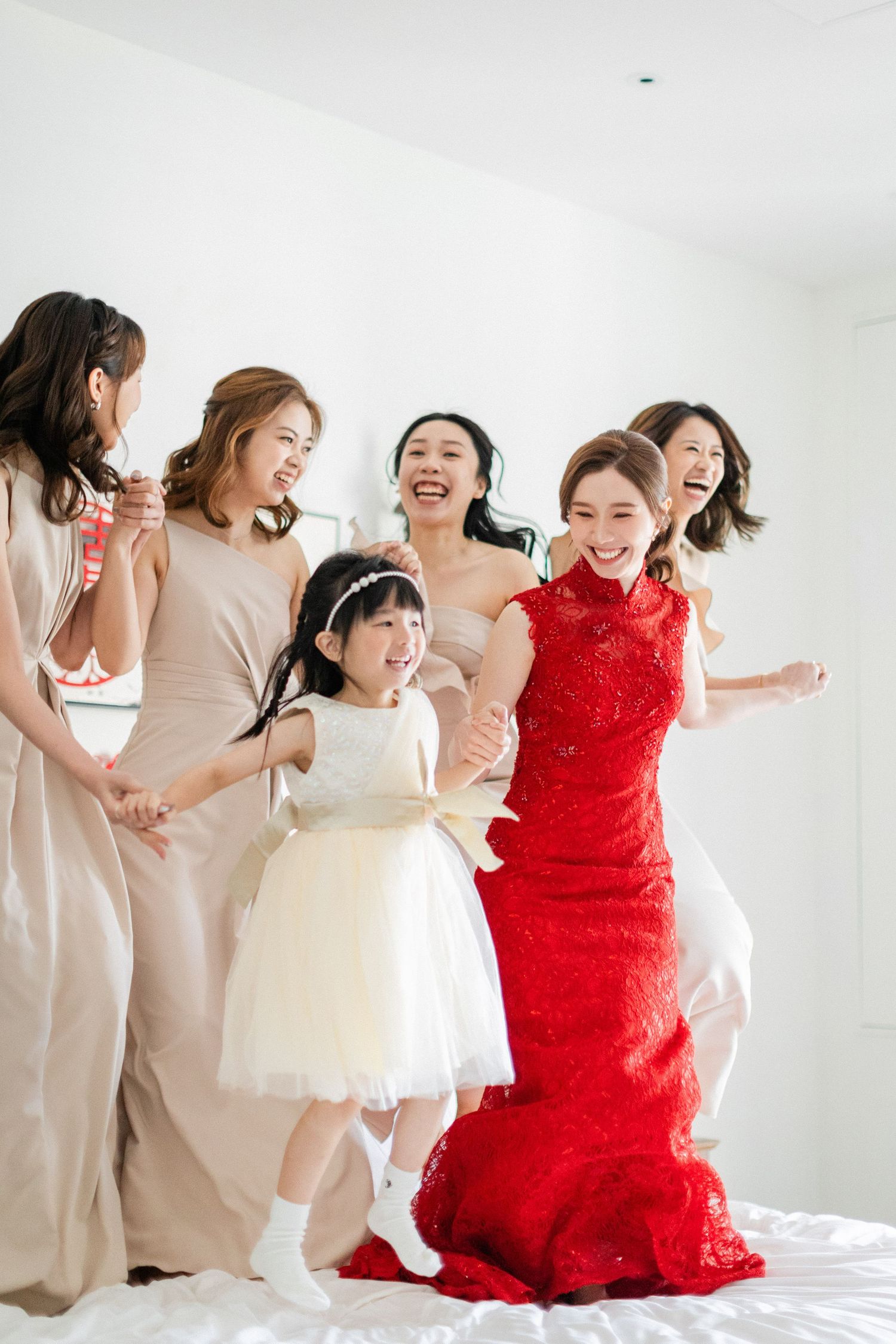A bride in a red Chinese qipao jumps excitedly on a bed with bridesmaids and a flower girl in a white dress.