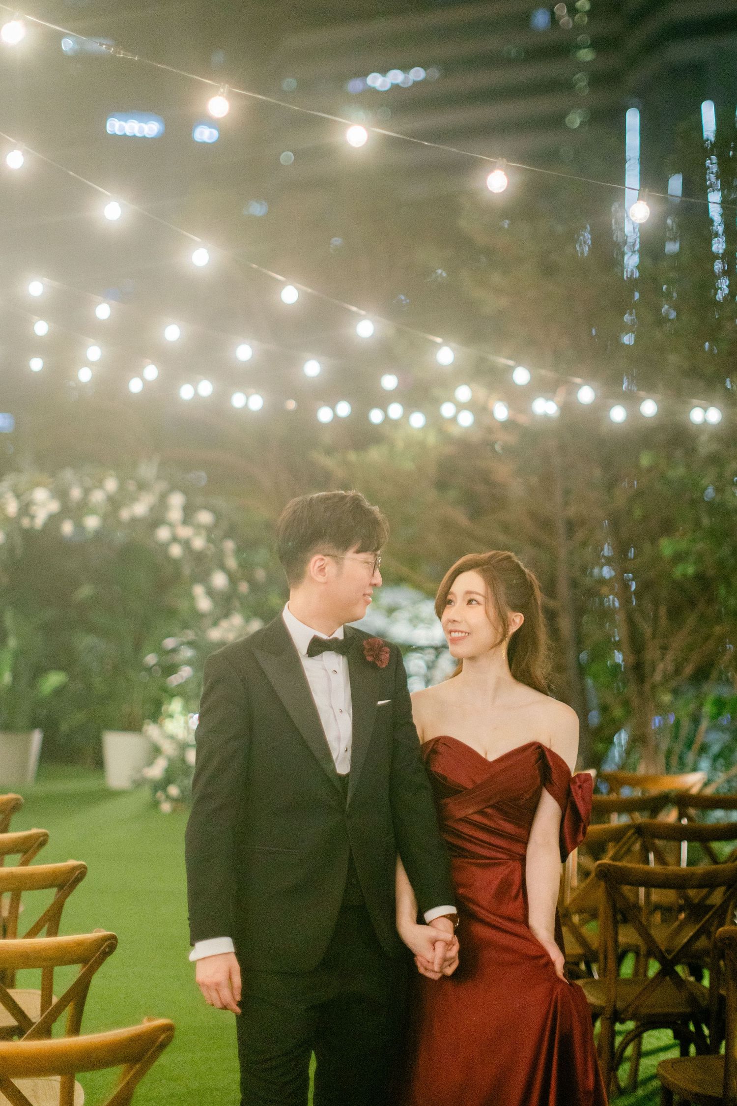 Couple in elegant evening attire hold hands under warm string lights at a romantic indoor wedding venue.