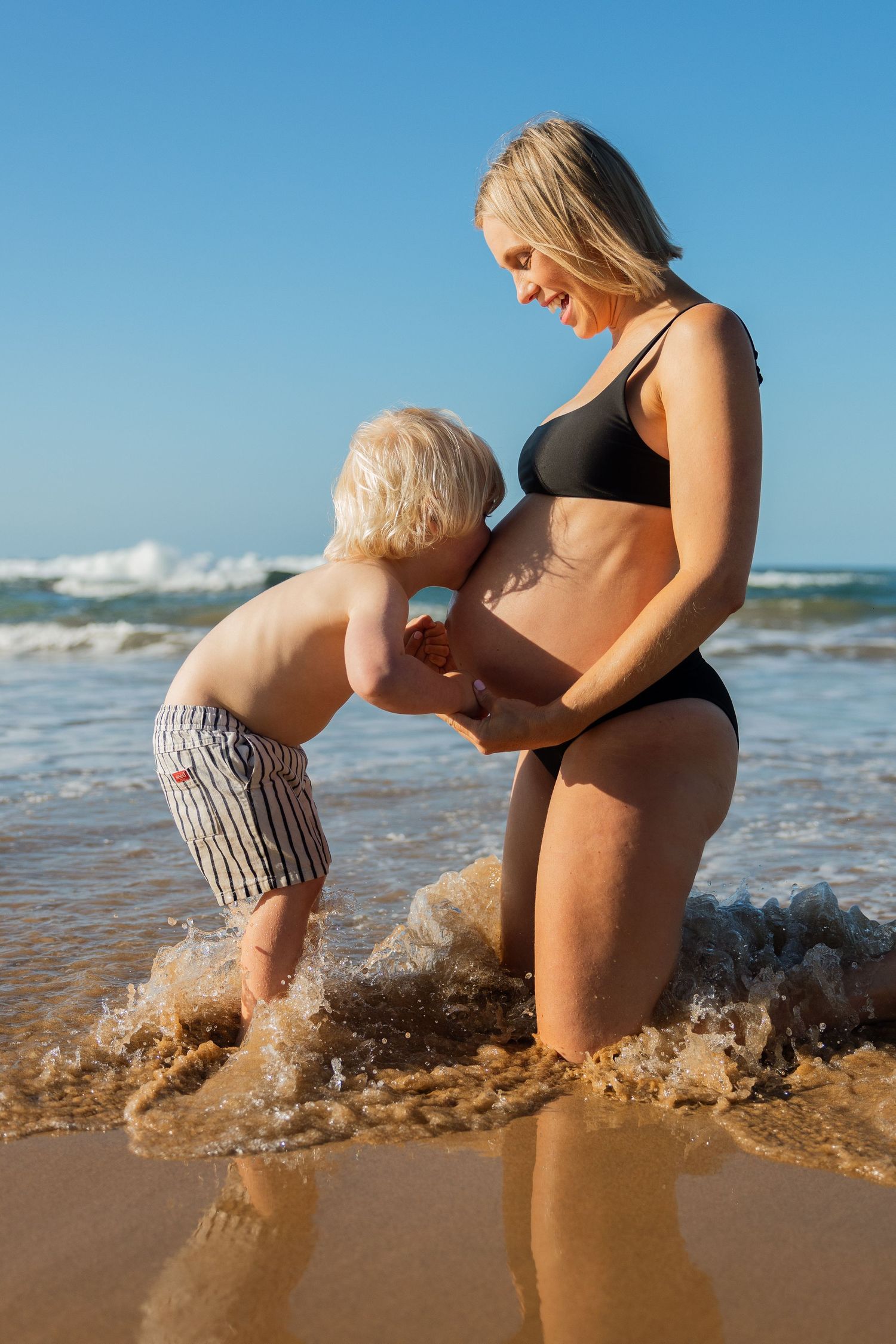 A tender moment between a small child and an expectant mother at the beach as waves crash behind them.