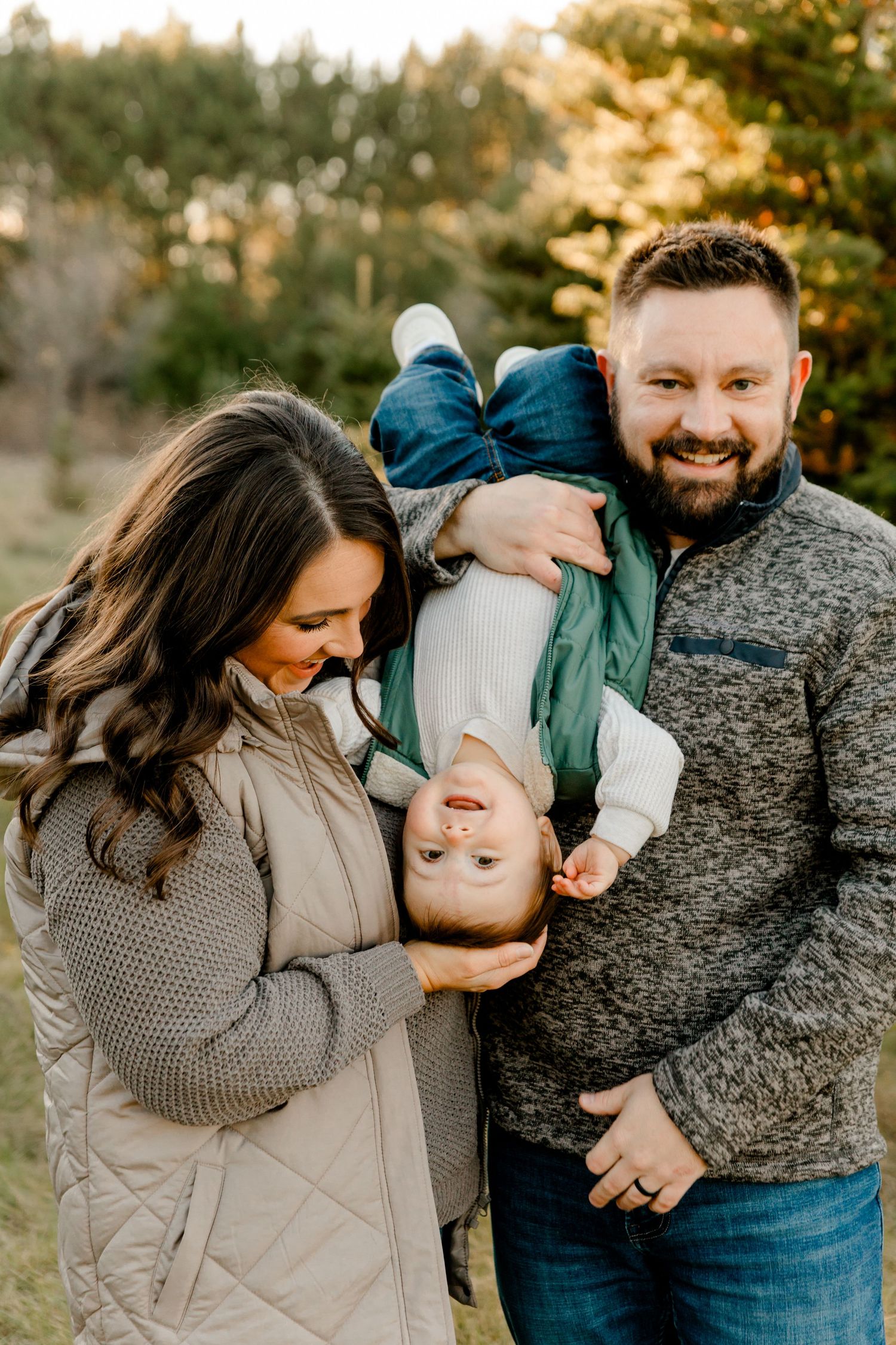 Laughing family holding baby upside down outdoors at a Christmas tree farm during fall.