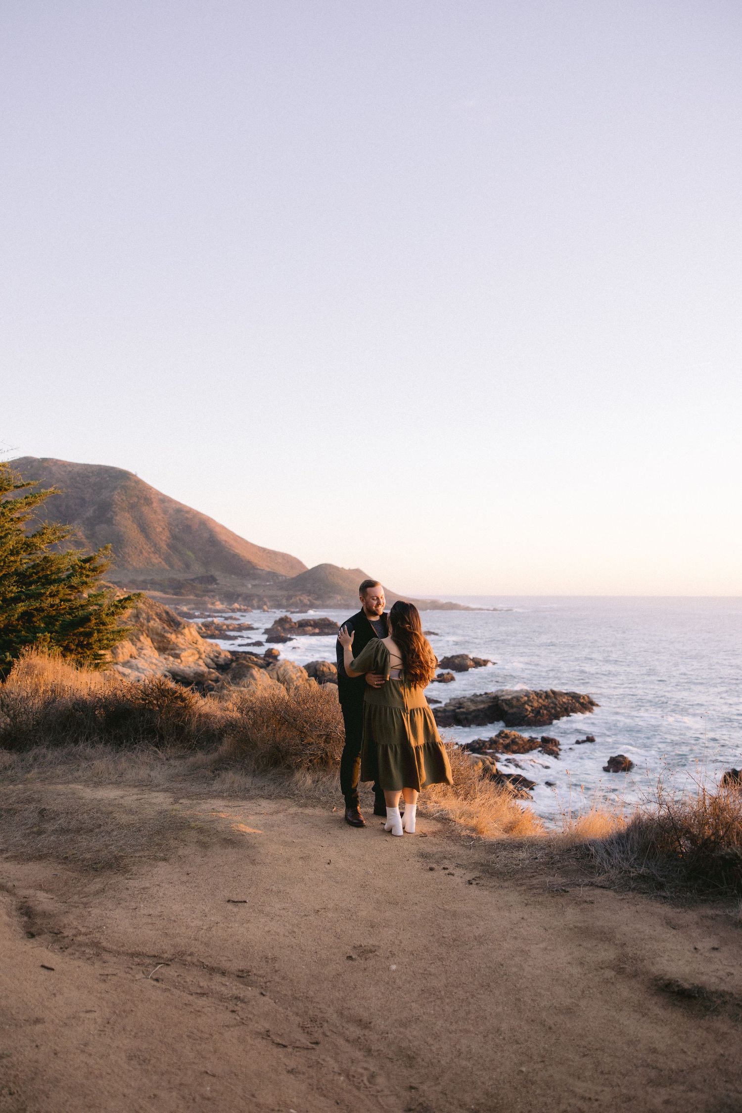 Two people standing together on a coastal cliff overlooking the rocky Pacific Ocean shoreline during golden hour.