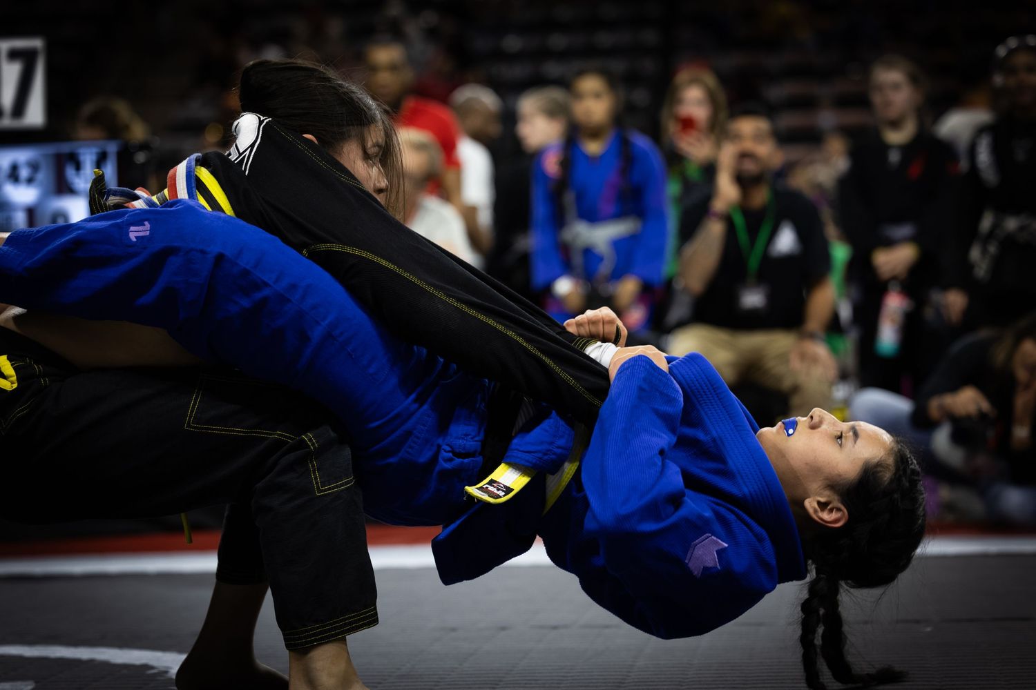 Dramatic action shot of Brazilian Jiu-Jitsu competitors grappling in dark arena setting.