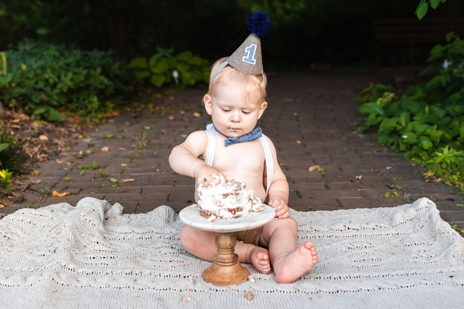 Baby in party hat sits on blanket with small birthday cake during outdoor celebration.