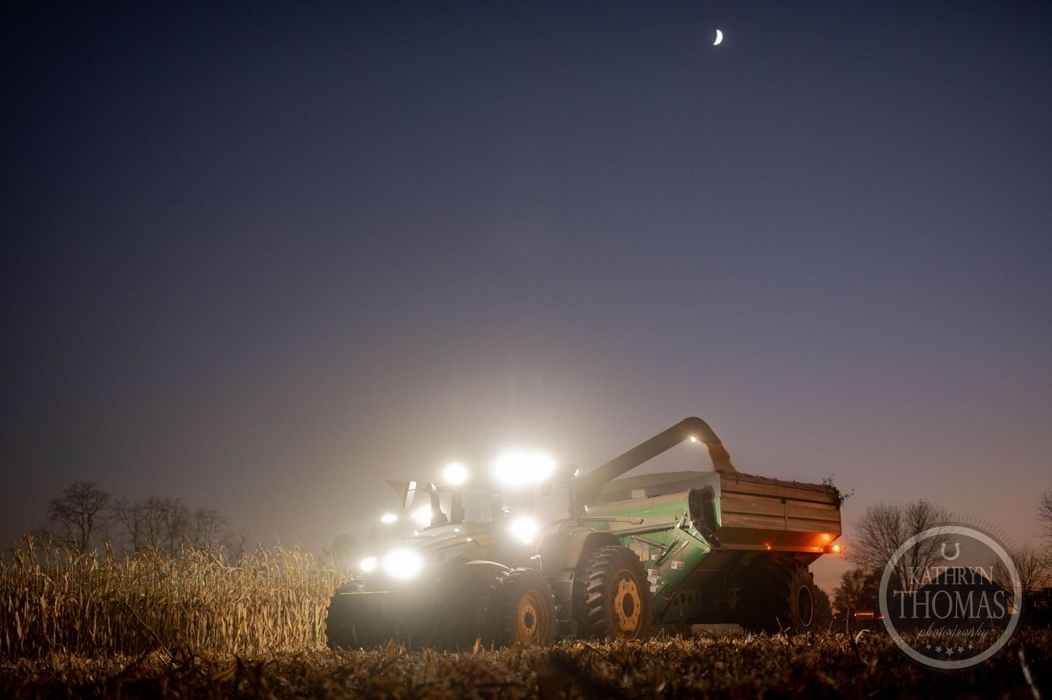 Agricultural machinery harvesting under moonlight with grain cart visible in field.