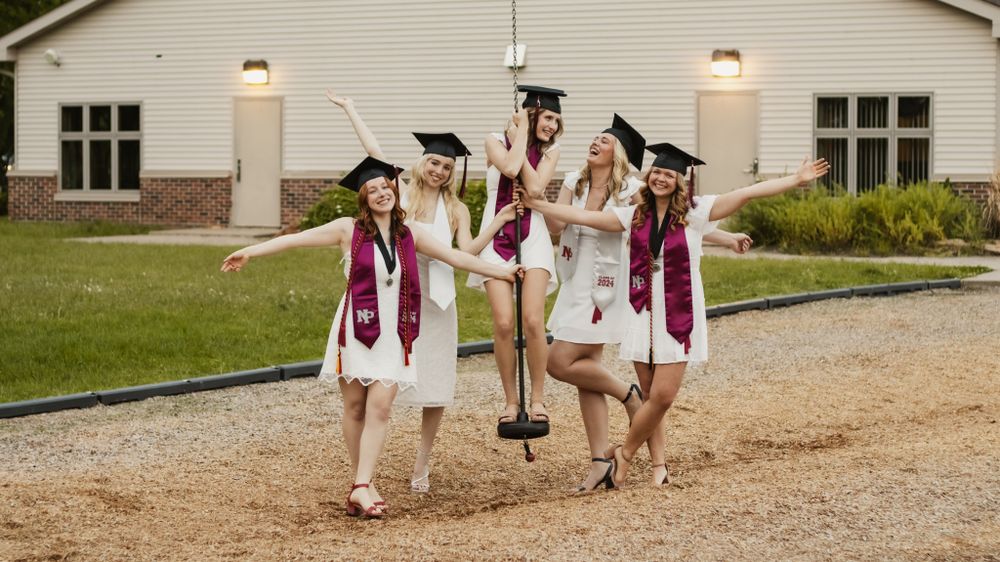 Graduates in white gowns and maroon stoles celebrating together outdoors.
