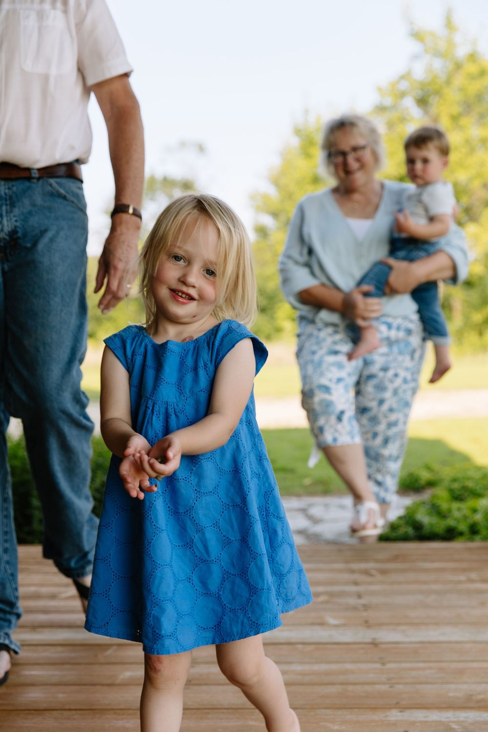 A child in a blue dress spins and dances playfully on a wooden deck during a sunny day outdoors.