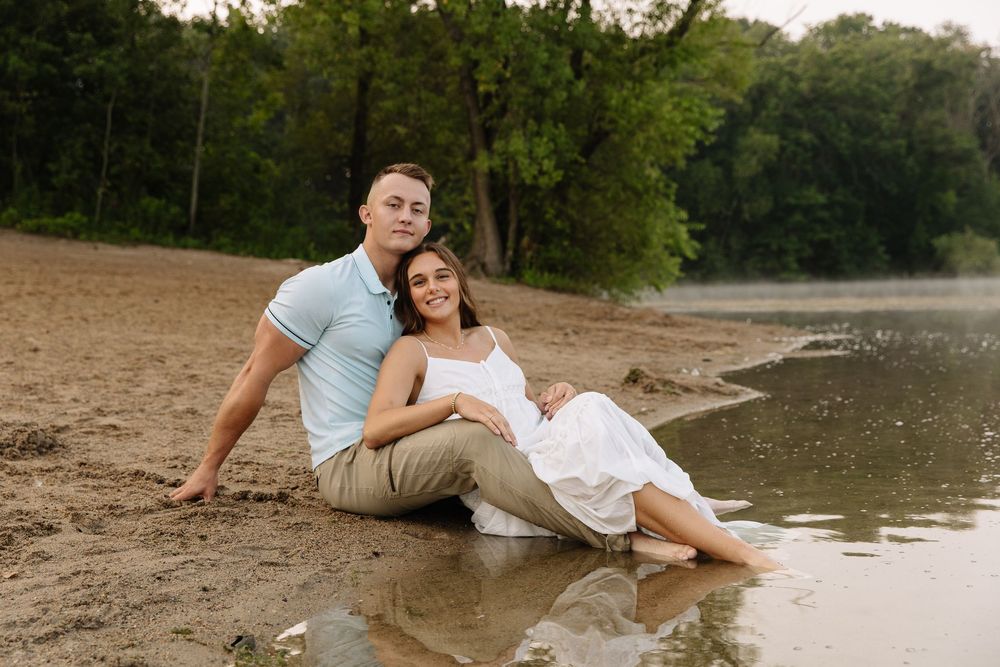 A couple shares a romantic moment on a sandy beach near a lake during golden hour.