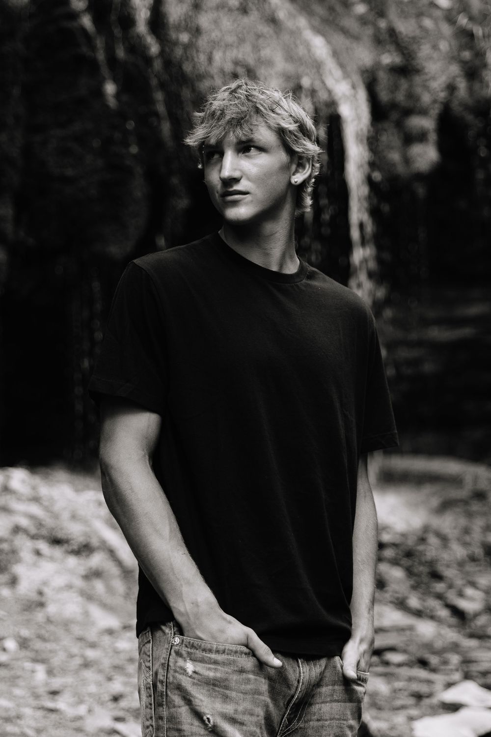 A person in a black t-shirt and jeans poses casually against a natural outdoor backdrop in black and white photography.
