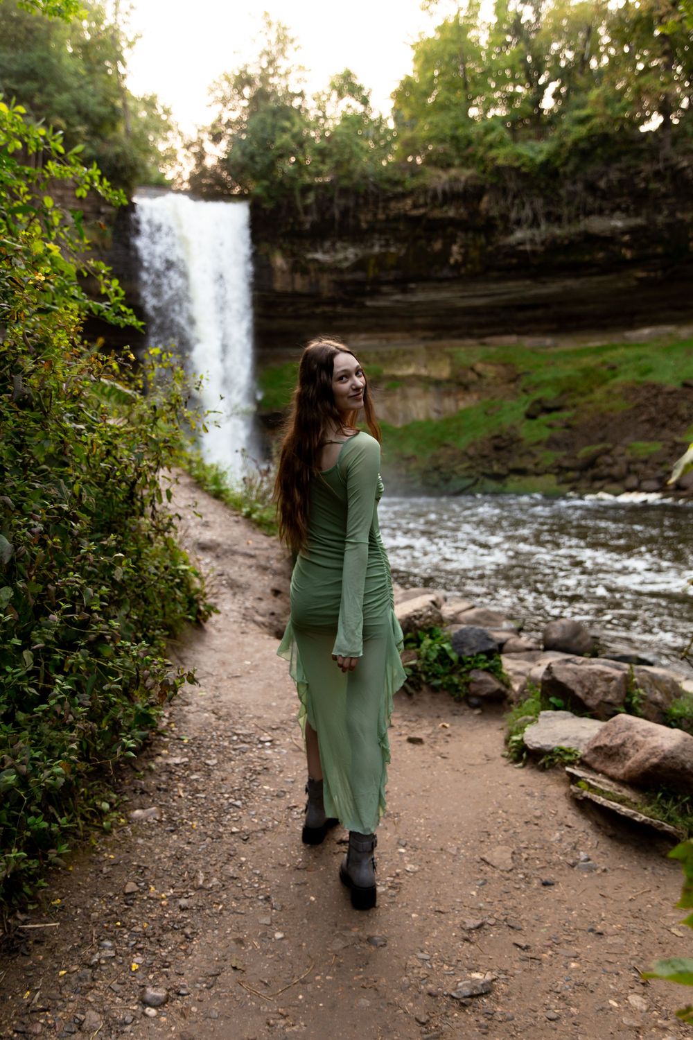 A person in a green sweater stands on a dirt path near a scenic waterfall in a wooded natural setting.