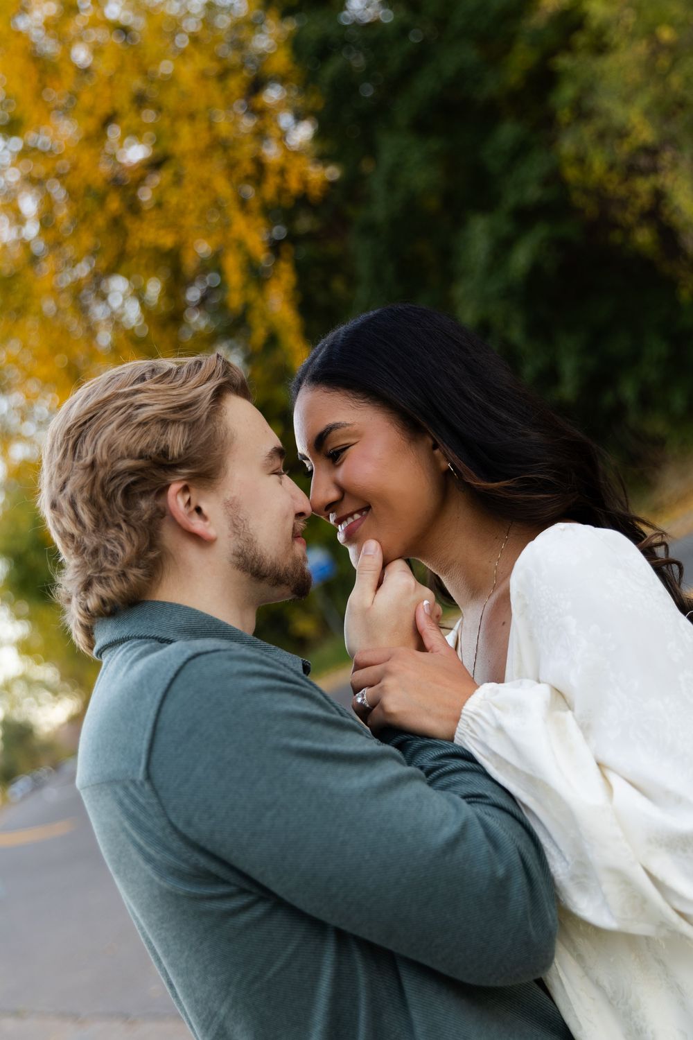 A romantic couple in autumn shares an intimate moment wearing white and green sweaters against golden fall foliage.