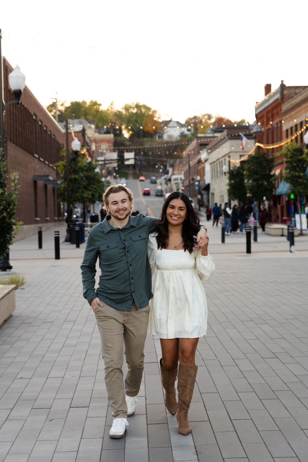 Happy couple poses playfully on a brick paved street at sunset with shops and buildings in the background.