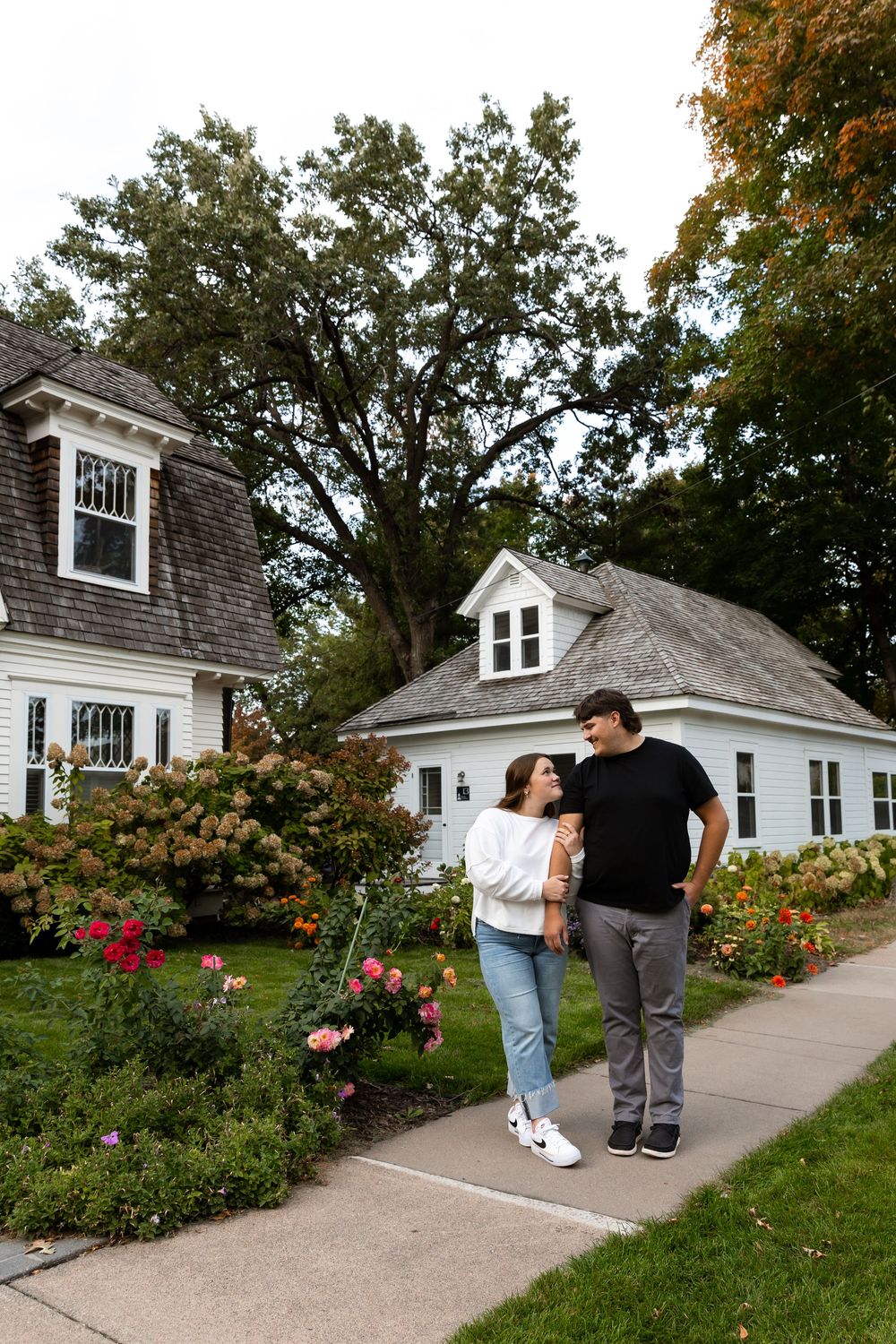 A couple shares a romantic moment walking along a garden path with blooming flowers and white houses in the background.
