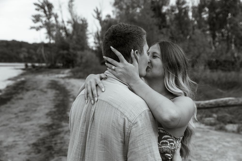 A romantic black and white photo shows a couple sharing an intimate kiss on a rural path surrounded by trees.