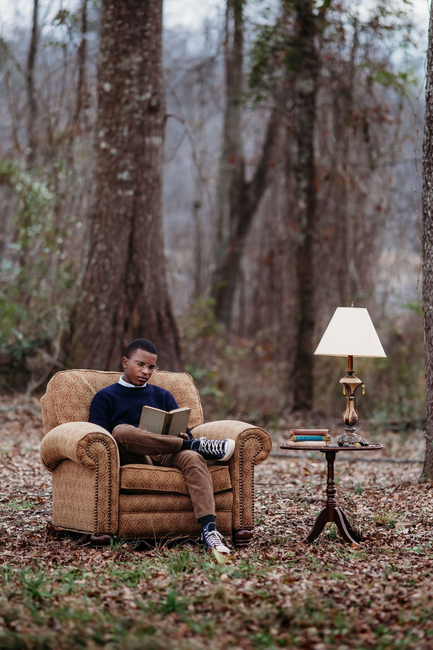 A cozy armchair and reading lamp setup in an autumn forest with fallen leaves on the ground.