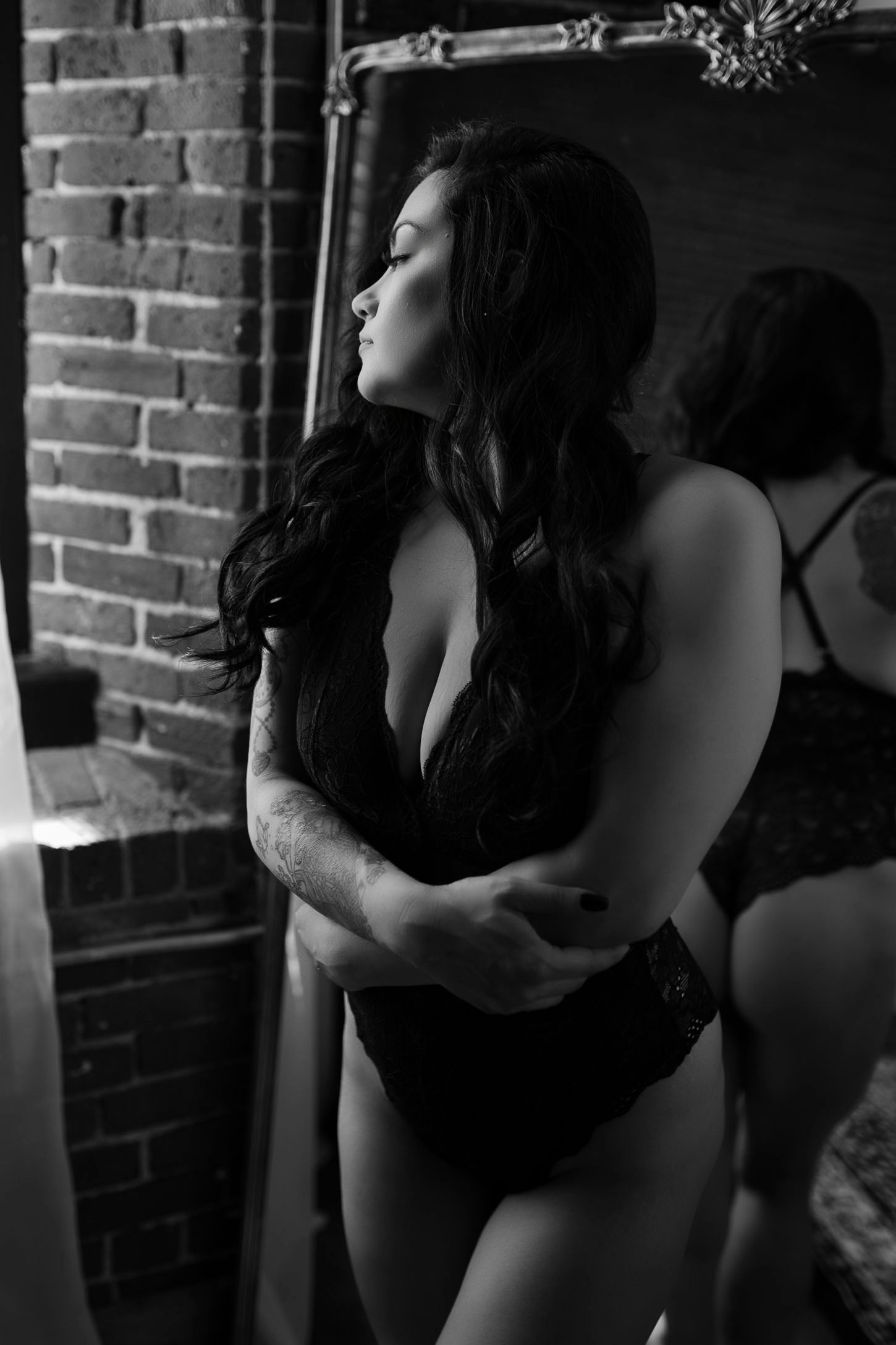 Artistic black and white boudoir photo of figure in lingerie posed against exposed brick wall in dramatic lighting.