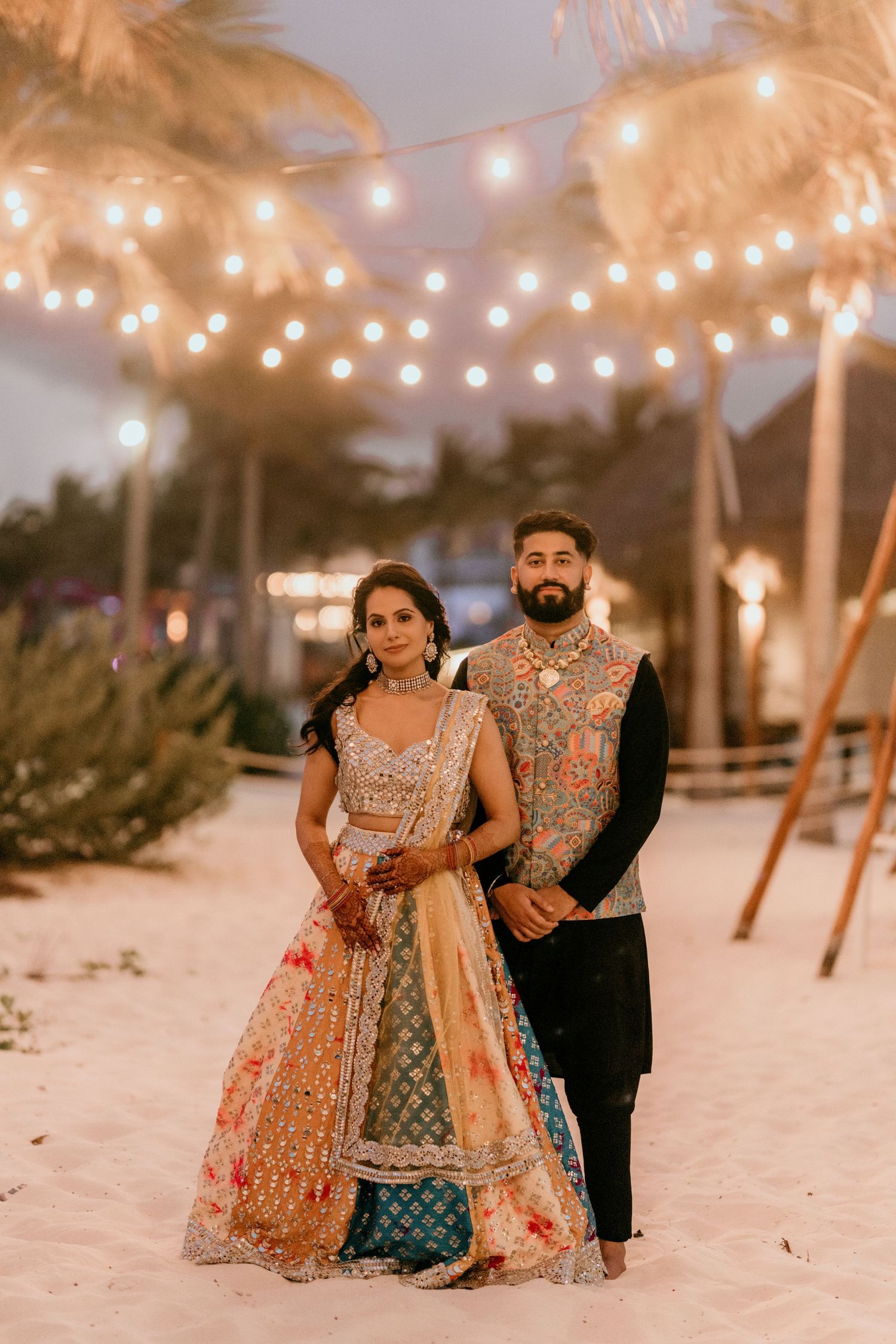 A couple in ornate traditional Indian wedding attire poses beneath string lights on a sandy beach at dusk.