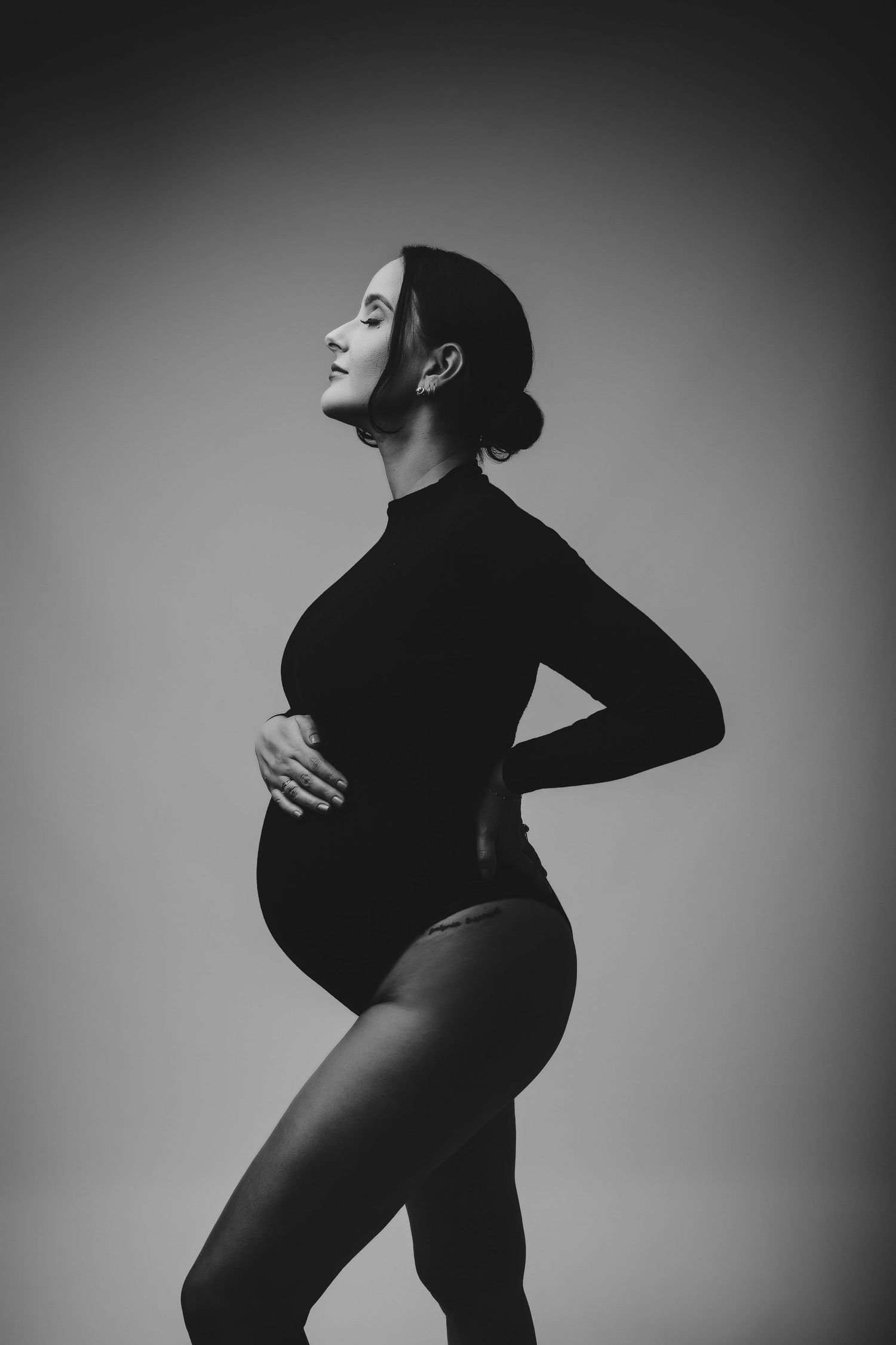 Moody maternity portrait of pregnant woman in black sweater and jeans against dark background.