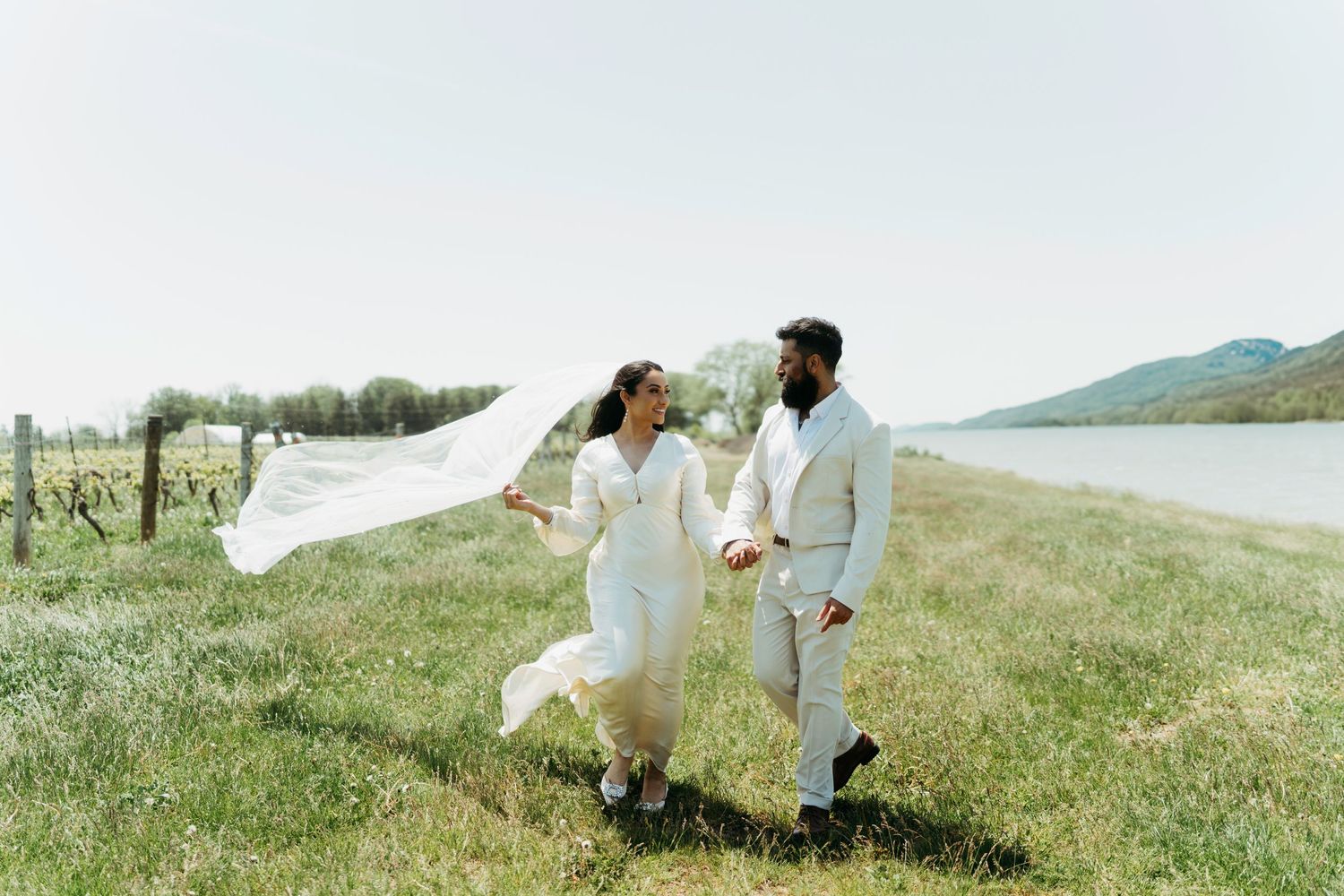 A couple in white wedding attire runs playfully through a grassy field near a lake with the bride's veil flowing in the wind.