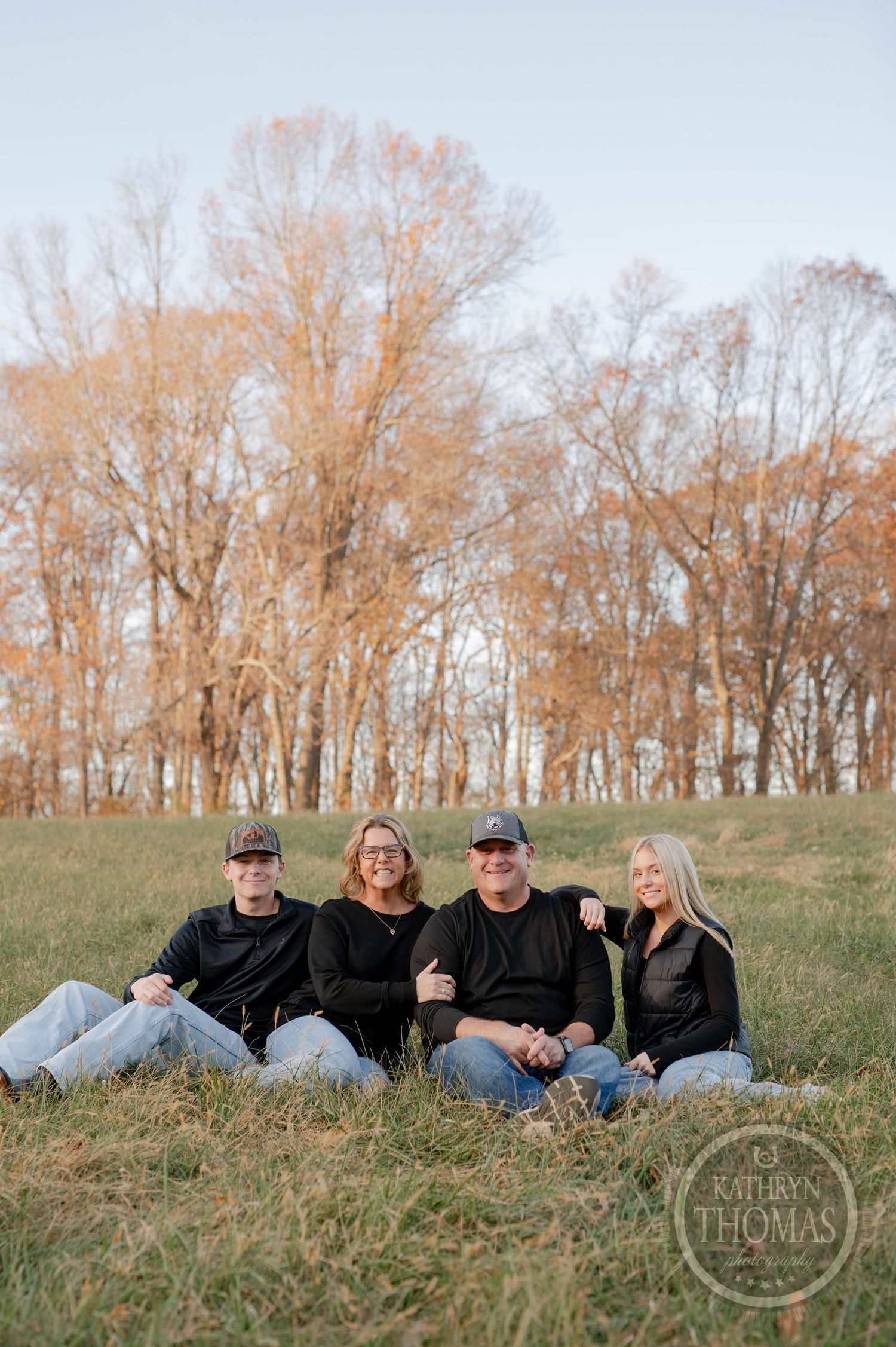 Group portrait of family members relaxing on grass against autumn foliage backdrop.