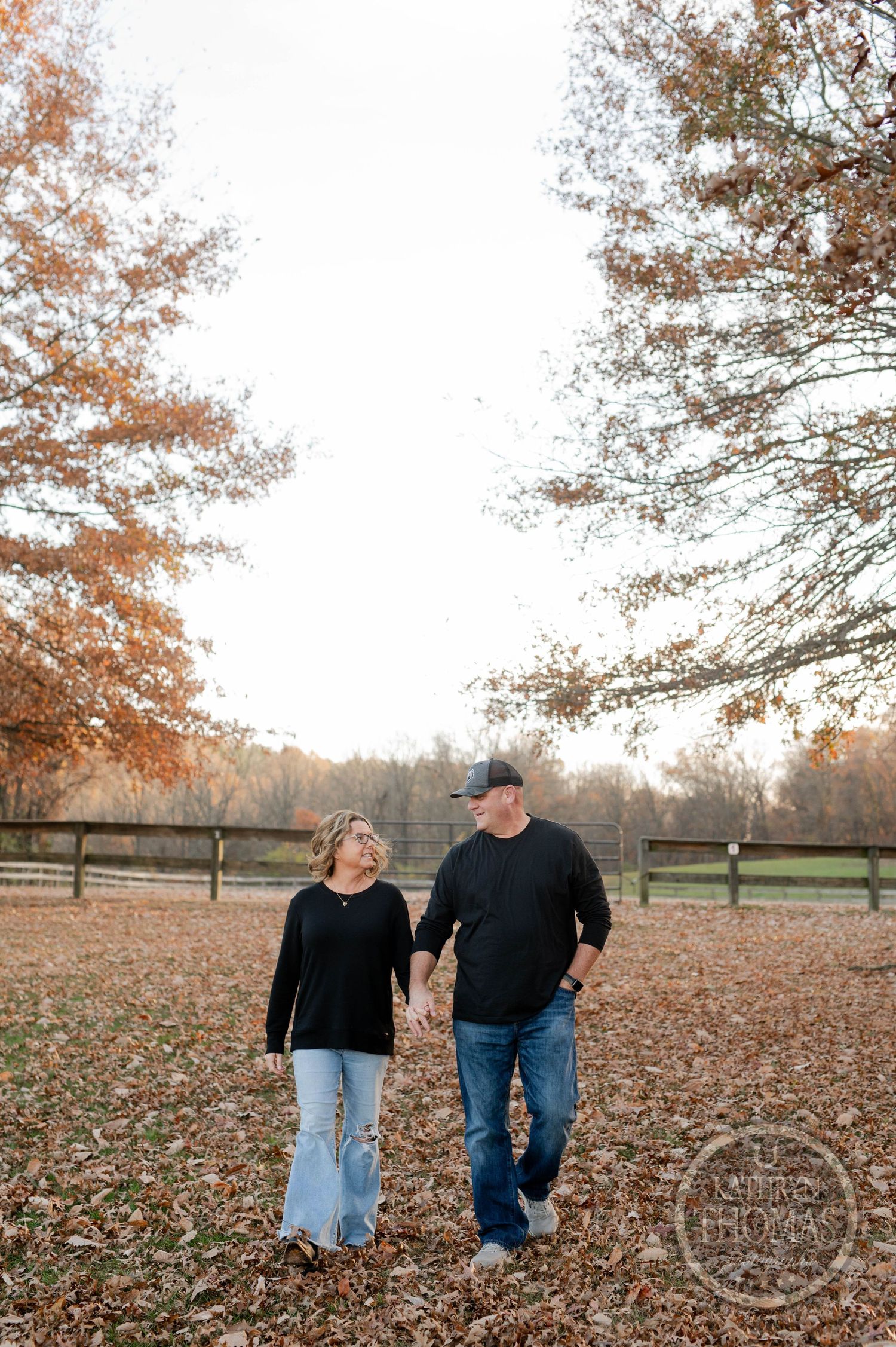 Couple takes romantic walk through fallen leaves with wooden fence and autumn trees in background.