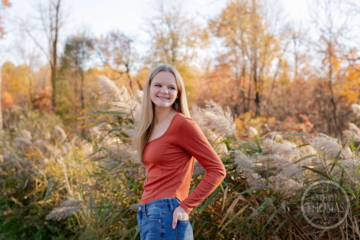Person in rustic orange top smiling against autumn foliage and golden light.