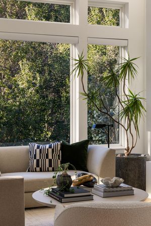 Sunlit living room corner with tall windows, indoor plants, and modern cream furniture.