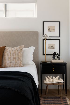 Minimalist bedroom corner with black nightstand, framed artwork, and textured throw pillows.