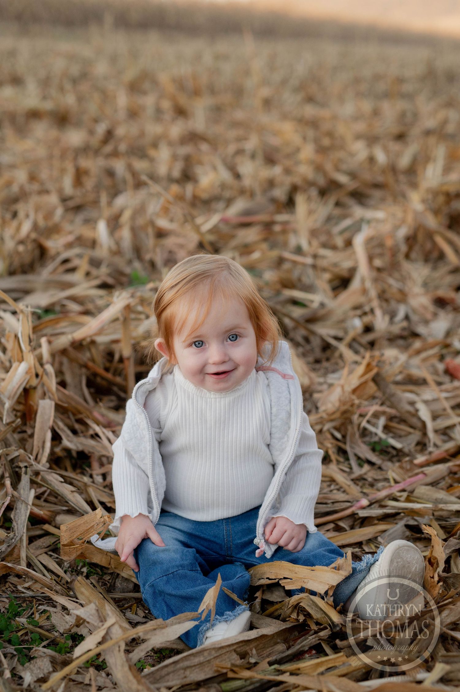Small child in white sweater sits among dried corn stalks in a harvested field.