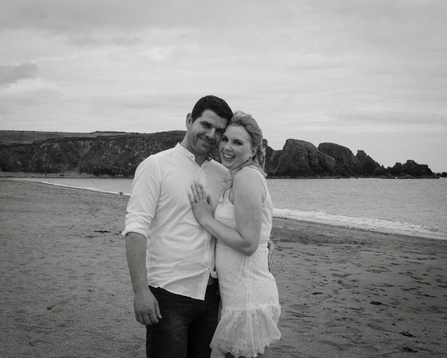 Black and white beach photo of two people embracing on a sandy shore with rocky cliffs in the background.