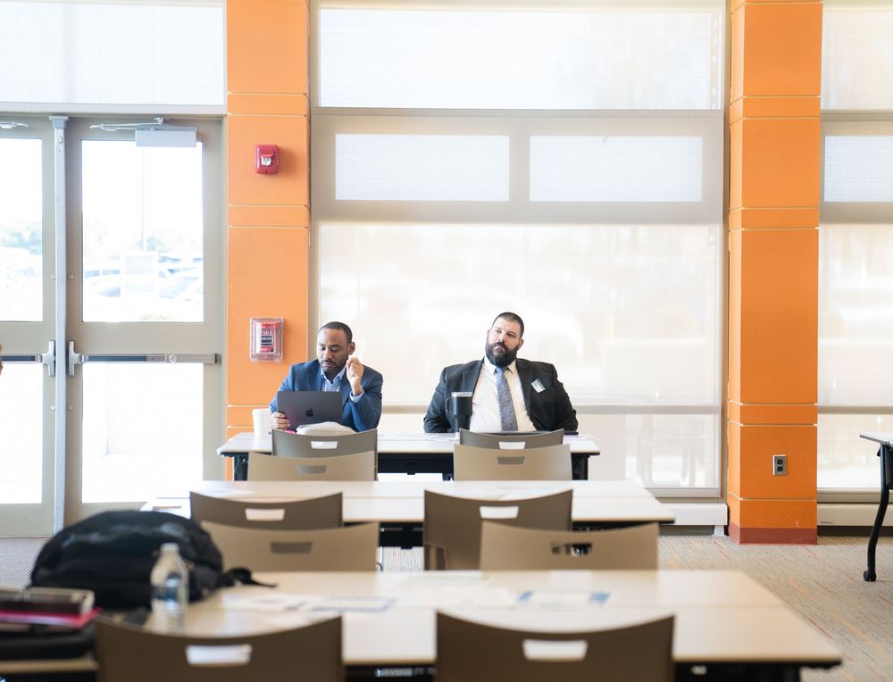 Two professionals sit at desks in a bright modern classroom with orange walls and empty student seating.
