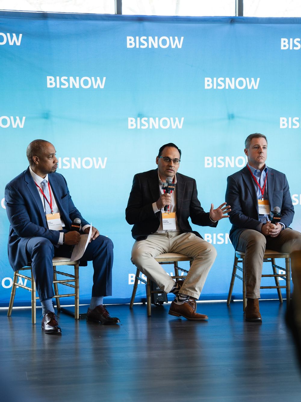 Three professionals sit on chairs at a Bisnow conference panel discussion, with the middle speaker holding a microphone.