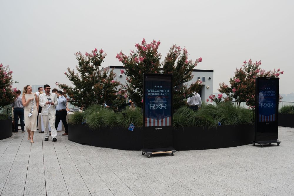 Rooftop terrace Bisnow event with guests mingling near lush pink flowering plants and welcome signage on a hazy day.