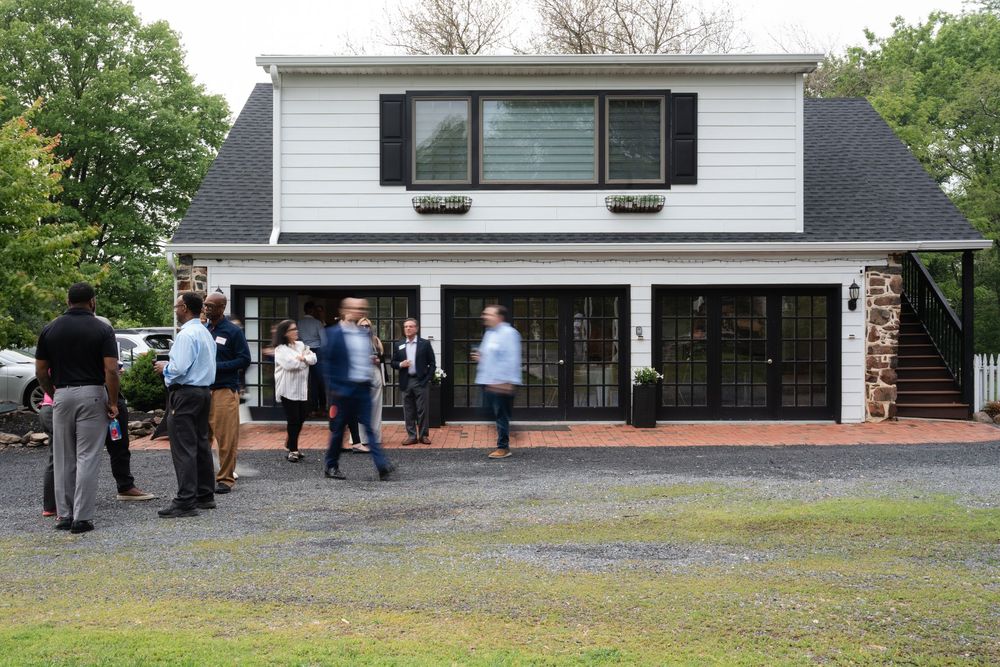 People gathering outside a white two-story building with black shutters and stone accents on a cloudy day.